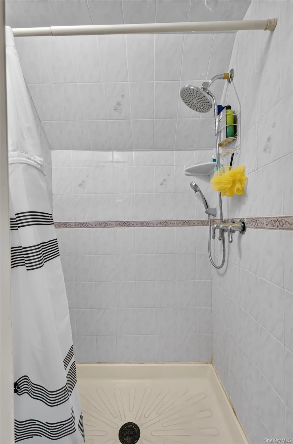 Shower stall featuring white ceramic tile, a decorative tile border, a circular shower head, and a handheld shower wand Shower stall featuring white ceramic tile, a decorative tile border, a circular shower head, and a handheld shower wand