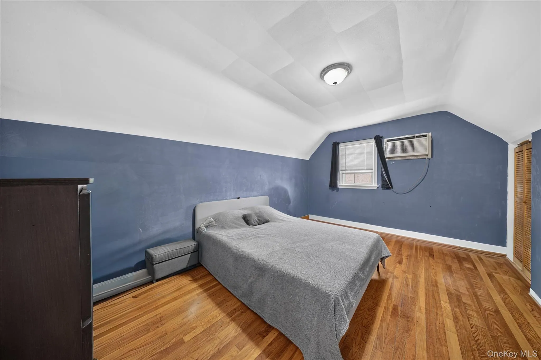 Room featuring wood-finish flooring, sloped ceilings, a window with blinds, and a built-in closet with louvered doors Room featuring wood-finish flooring, sloped ceilings, a window with blinds, and a built-in closet with louvered doors