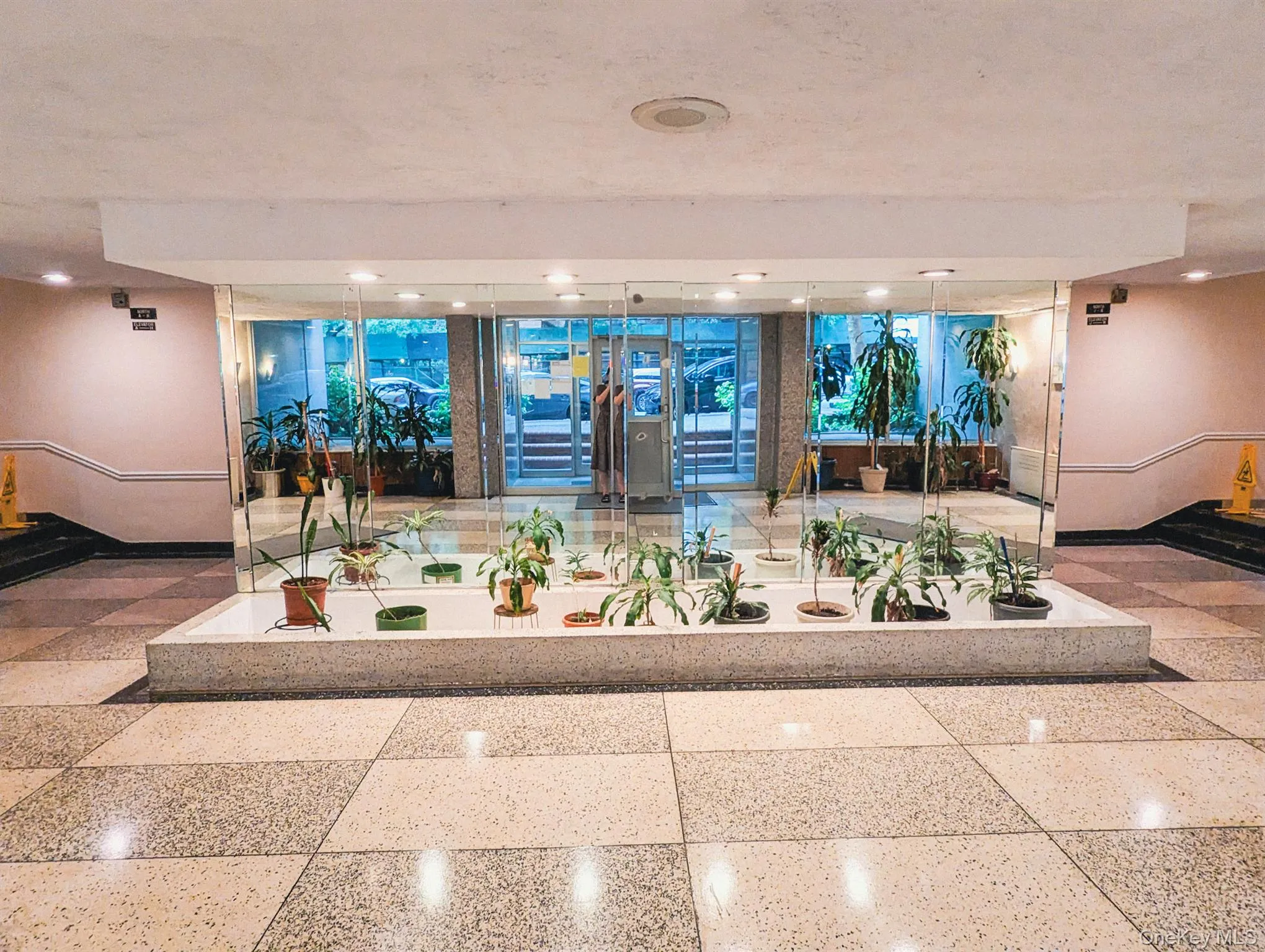 Lobby entrance featuring checkered tile flooring, a central planter box with assorted greenery, and a reflective glass wall Lobby entrance featuring checkered tile flooring, a central planter box with assorted greenery, and a reflective glass wall