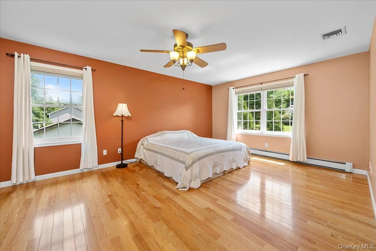 Spacious room featuring wood-finish flooring, two large windows, a ceiling fan with integrated lighting, and baseboard heating. Spacious room featuring wood-finish flooring, two large windows, a ceiling fan with integrated lighting, and baseboard heating.