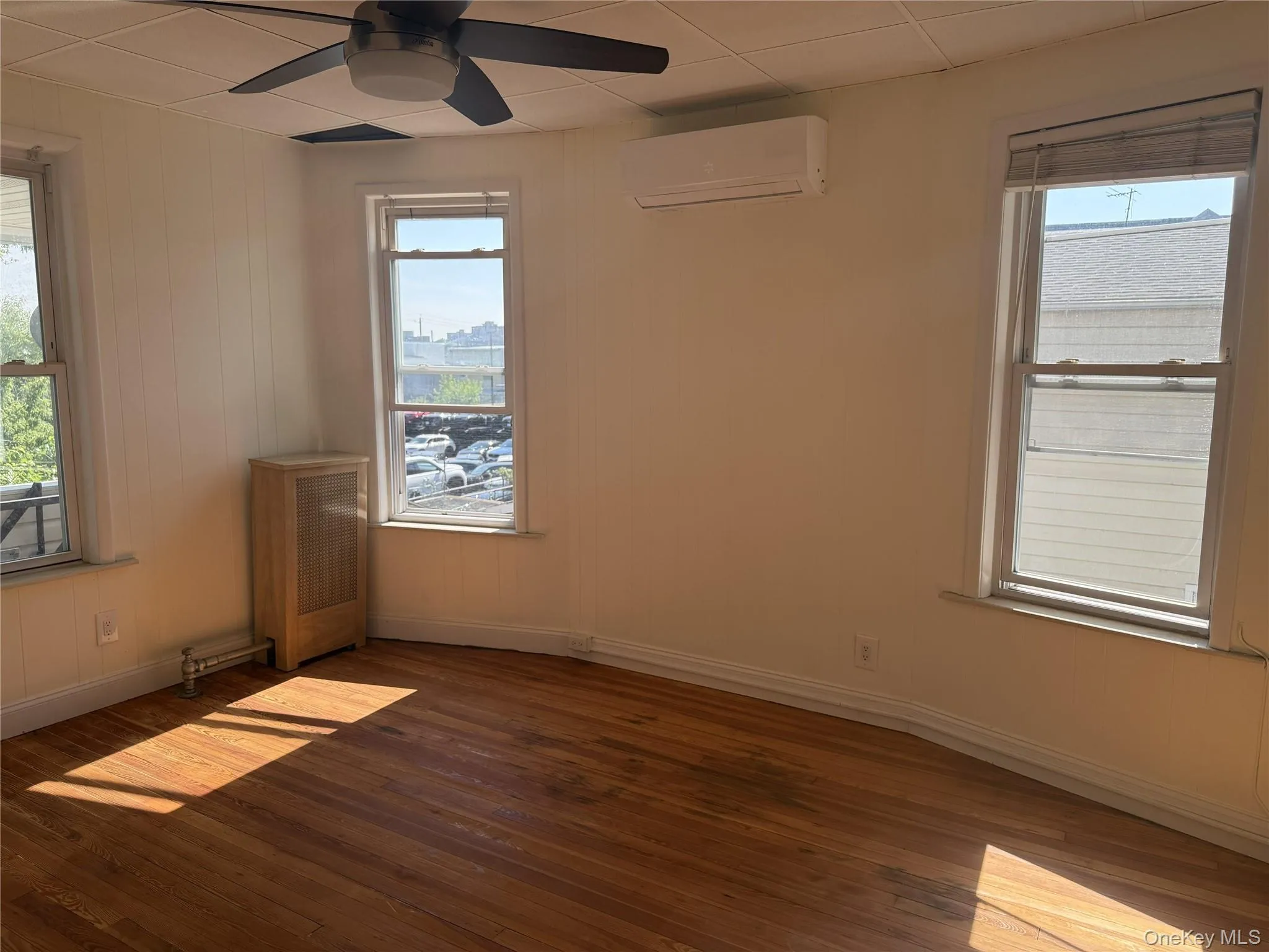 Room featuring wood-finish flooring, multiple windows, a ceiling fan with integrated lighting, a wall-mounted climate control unit, and white baseboards Room featuring wood-finish flooring, multiple windows, a ceiling fan with integrated lighting, a wall-mounted climate control unit, and white baseboards
