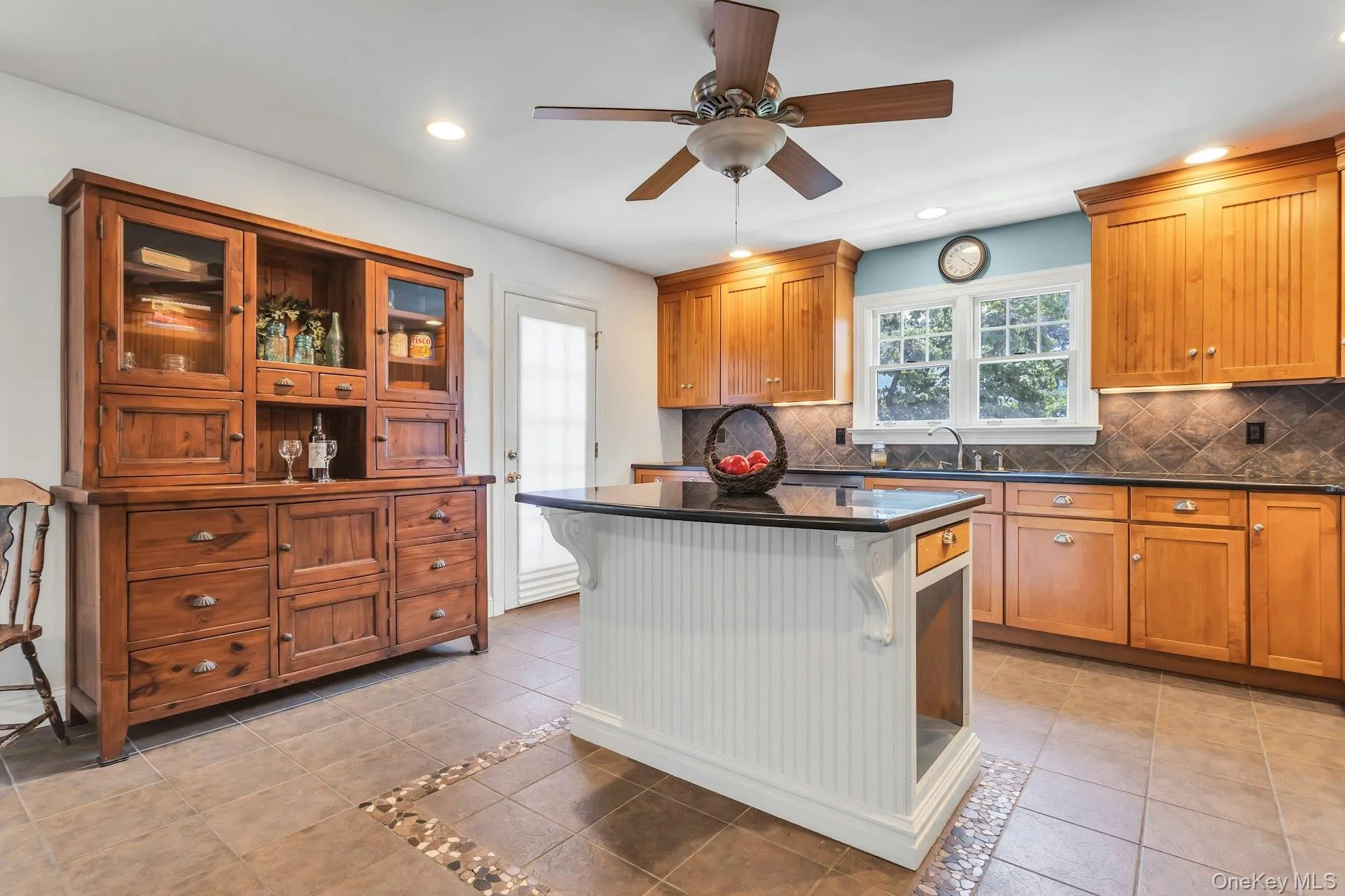 Kitchen featuring a central island with beadboard paneling and a black countertop, perimeter wood cabinetry, a tile backsplash, and a ceiling fan Kitchen featuring a central island with beadboard paneling and a black countertop, perimeter wood cabinetry, a tile backsplash, and a ceiling fan