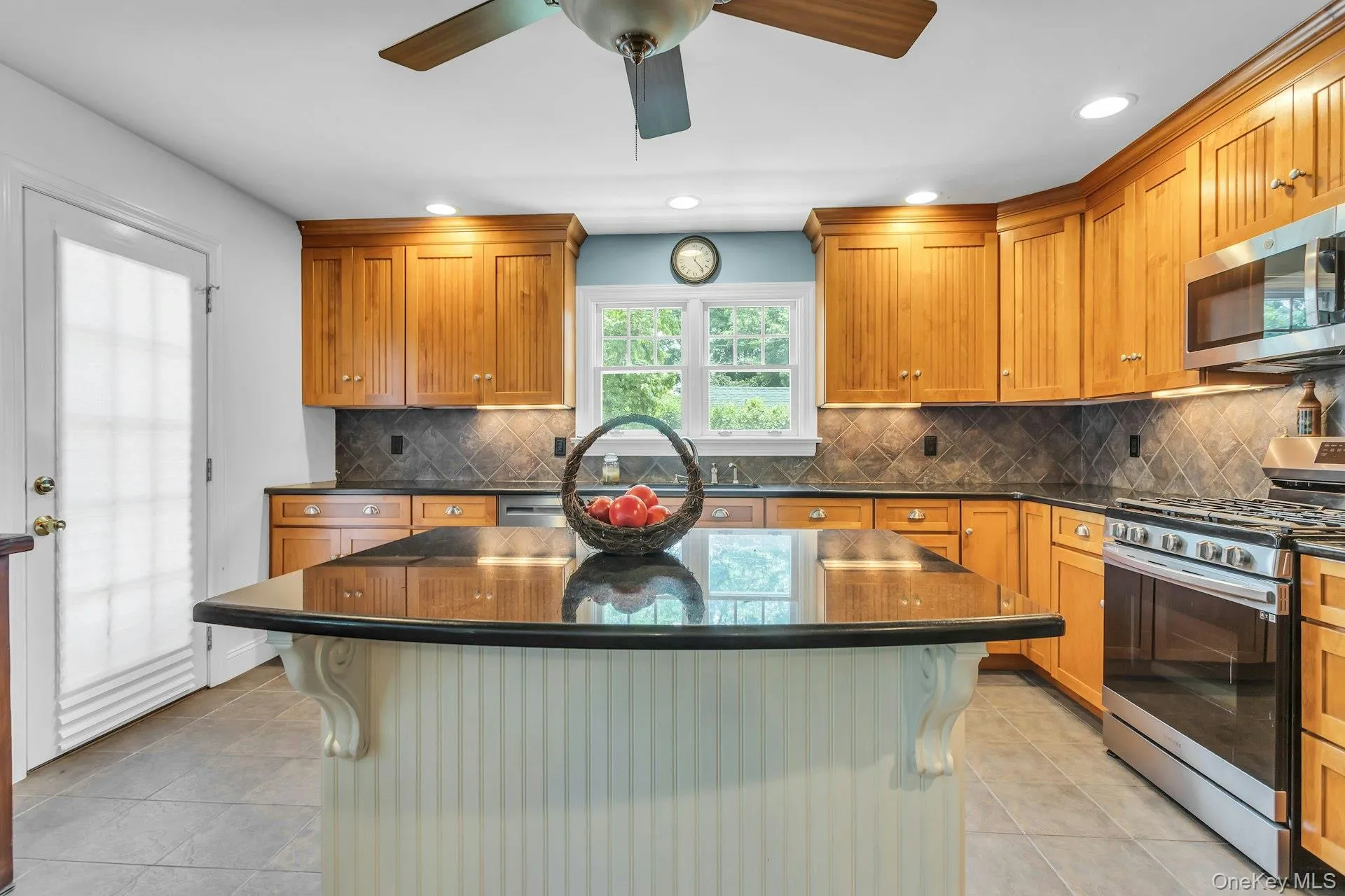 Kitchen featuring a central island with a dark countertop, light wood cabinetry, stainless steel appliances, a tile backsplash, and tile flooring Kitchen featuring a central island with a dark countertop, light wood cabinetry, stainless steel appliances, a tile backsplash, and tile flooring
