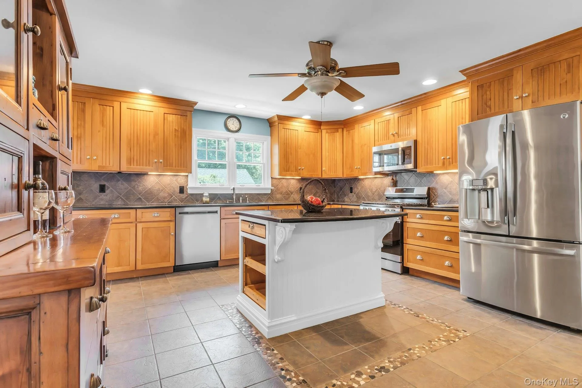 Kitchen featuring custom wood cabinetry, stainless steel appliances, a central island with a contrasting white base, a tiled backsplash, and recessed lighting Kitchen featuring custom wood cabinetry, stainless steel appliances, a central island with a contrasting white base, a tiled backsplash, and recessed lighting