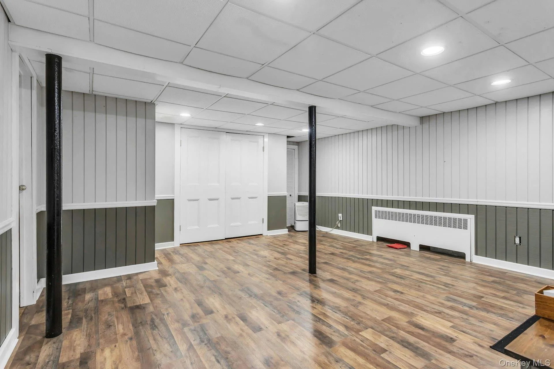 Spacious finished room featuring wood-finish flooring, two-tone vertical paneling, and a drop ceiling with recessed lighting Spacious finished room featuring wood-finish flooring, two-tone vertical paneling, and a drop ceiling with recessed lighting