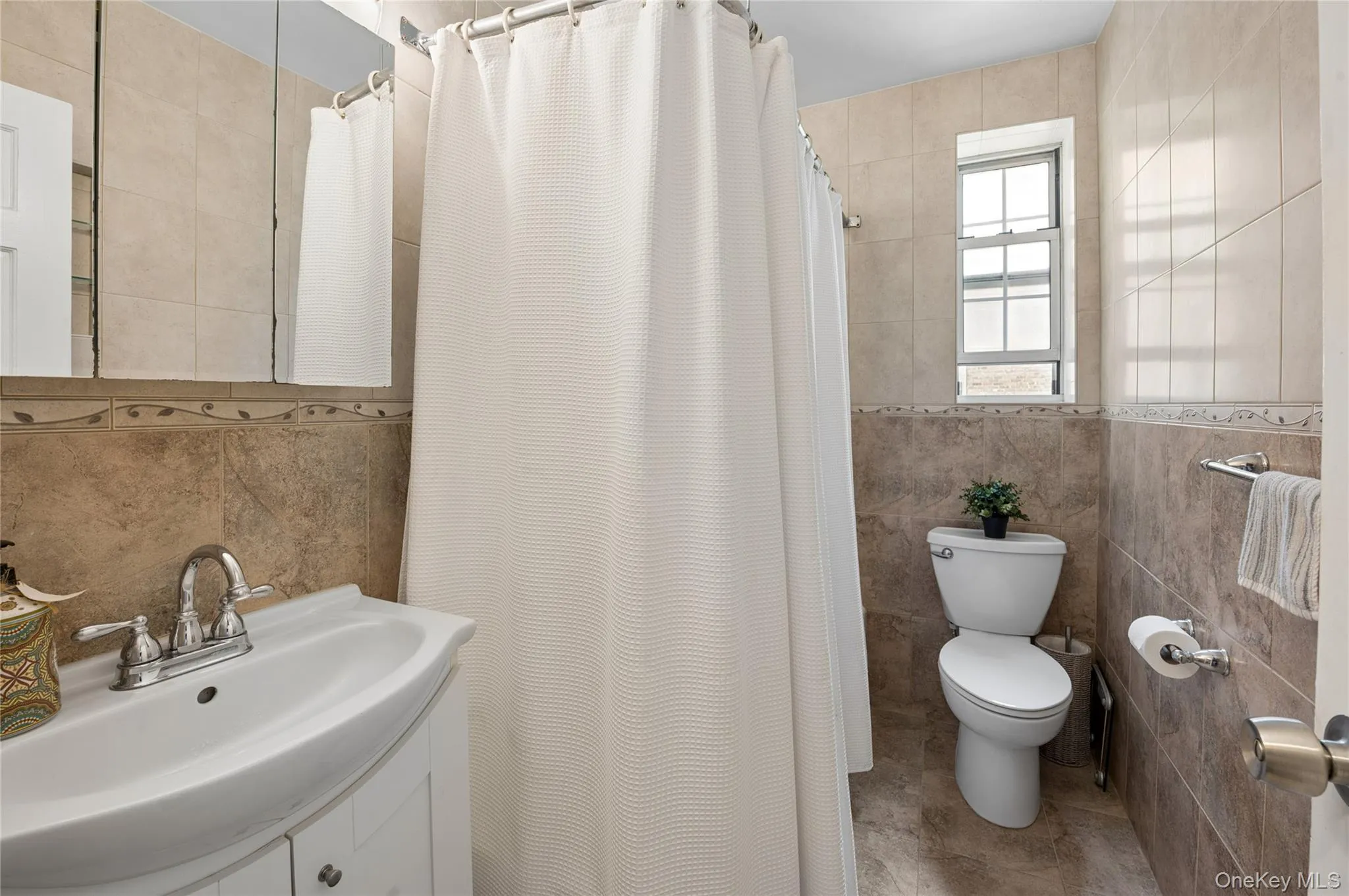 Bathroom featuring a vanity with a white ceramic sink, chrome faucet, tiled walls with decorative trim, and a window for natural light Bathroom featuring a vanity with a white ceramic sink, chrome faucet, tiled walls with decorative trim, and a window for natural light