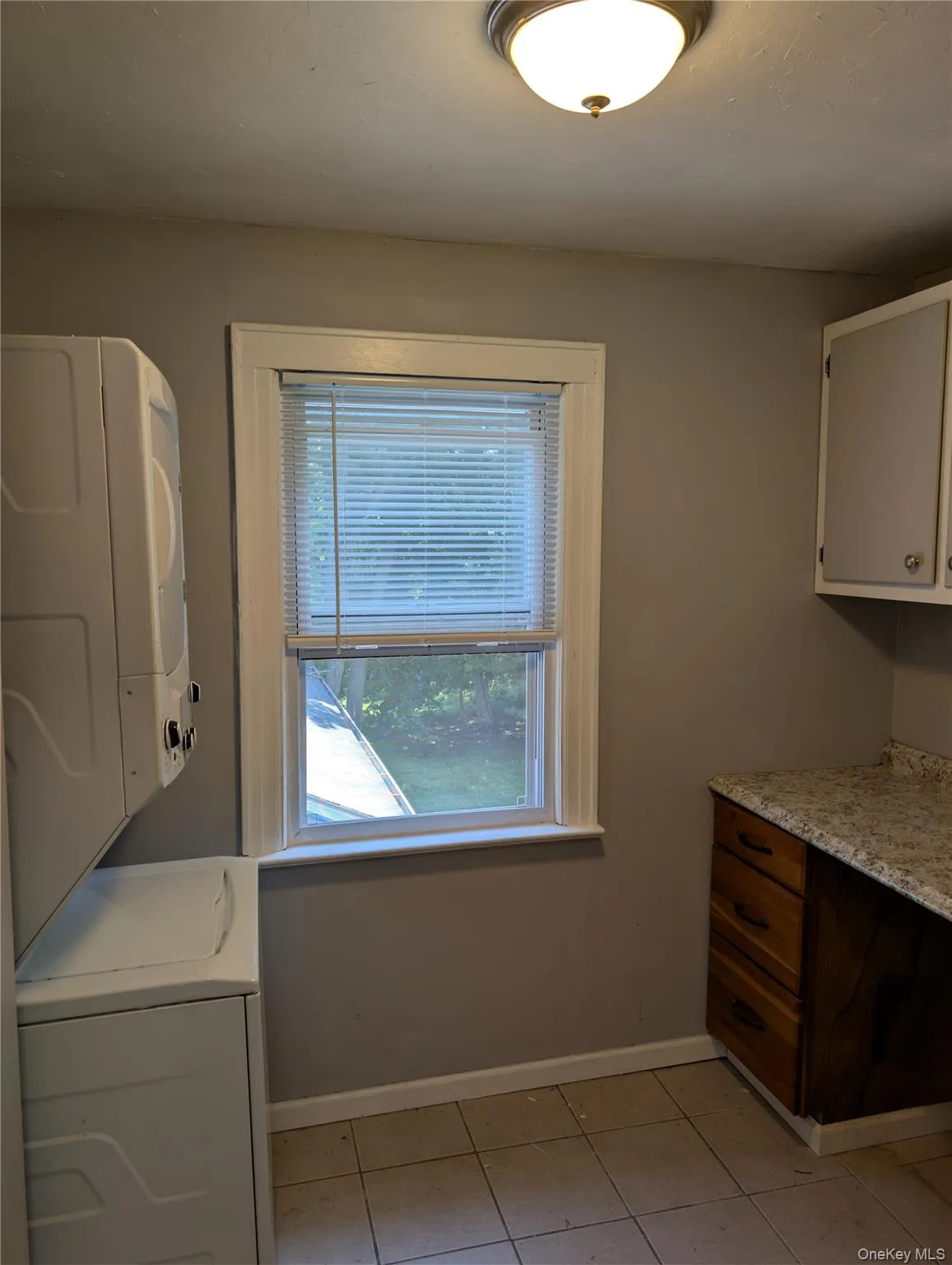 Laundry room features a window, overhead flush-mount light fixture, base cabinetry with a light-toned countertop and stackable washer/dryer combination Laundry room features a window, overhead flush-mount light fixture, base cabinetry with a light-toned countertop and stackable washer/dryer combination