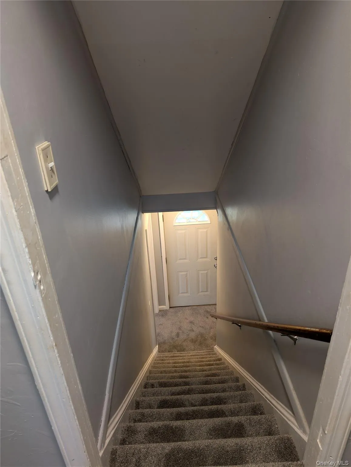 Carpeted staircase with light gray walls and a wooden handrail, leading to entrance to exterior, den or living room Carpeted staircase with light gray walls and a wooden handrail, leading to entrance to exterior, den or living room