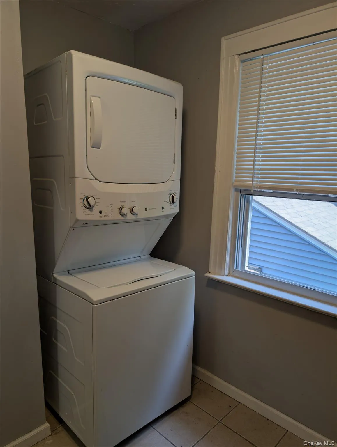 Dedicated laundry area features a stacked washer and dryer unit, tiled flooring, painted walls, and a window with blinds Dedicated laundry area features a stacked washer and dryer unit, tiled flooring, painted walls, and a window with blinds