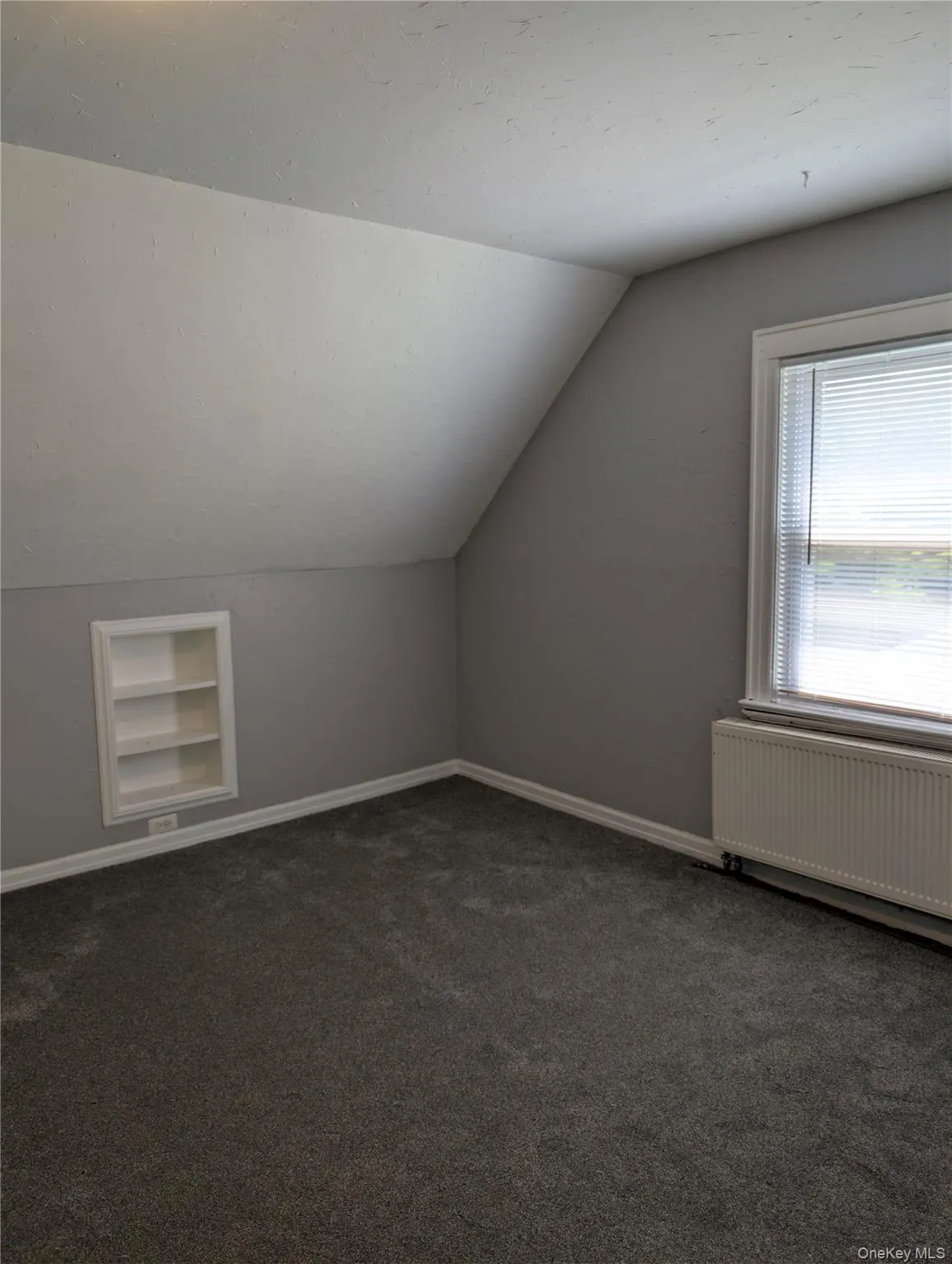Carpeted bedroom features sloped ceilings, a built-in shelving unit, and a window with blinds Carpeted bedroom features sloped ceilings, a built-in shelving unit, and a window with blinds