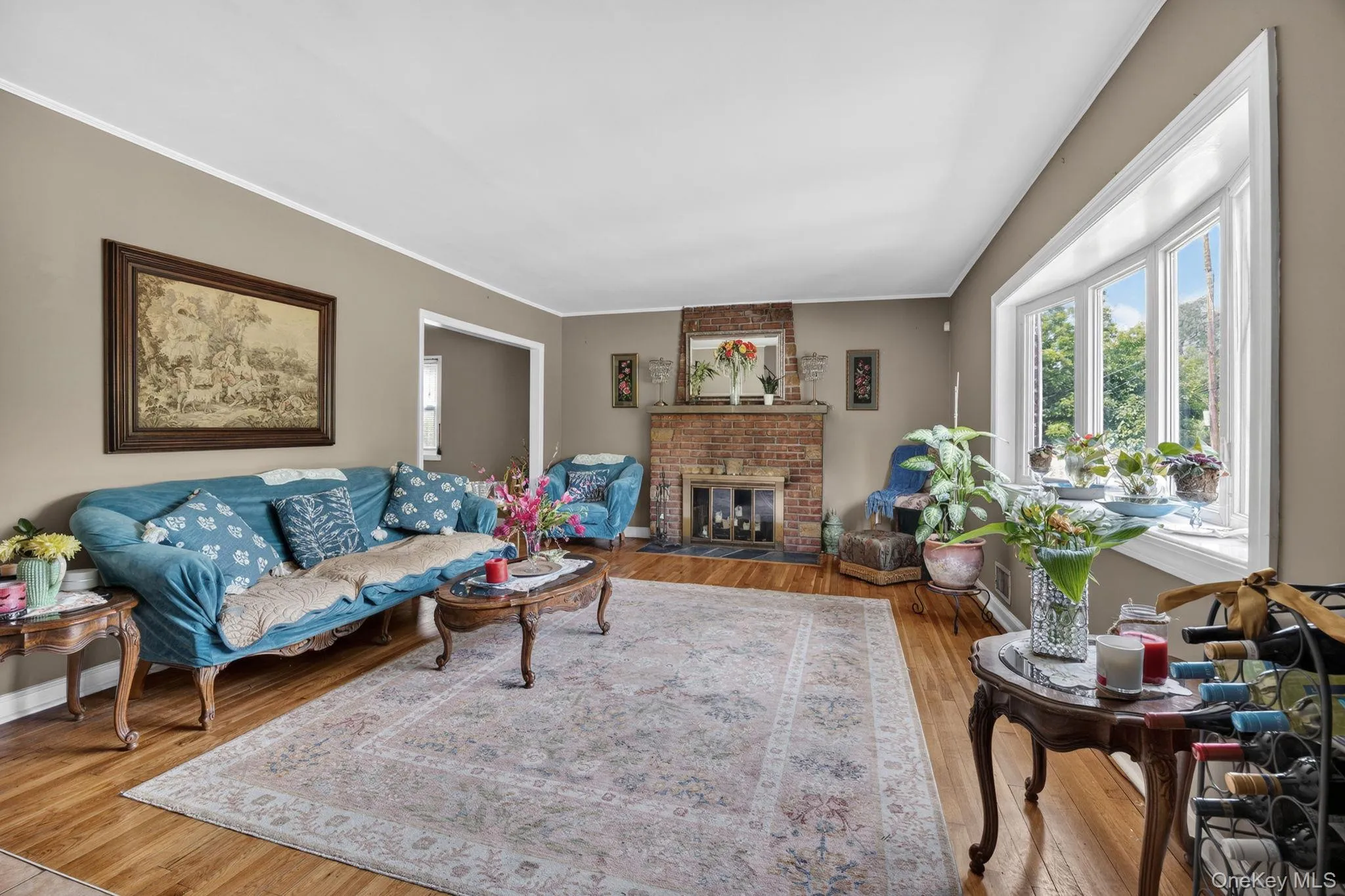 Living area featuring hardwood flooring, a brick fireplace with a decorative mantel, a large bay window, and coved ceilings with crown molding Living area featuring hardwood flooring, a brick fireplace with a decorative mantel, a large bay window, and coved ceilings with crown molding