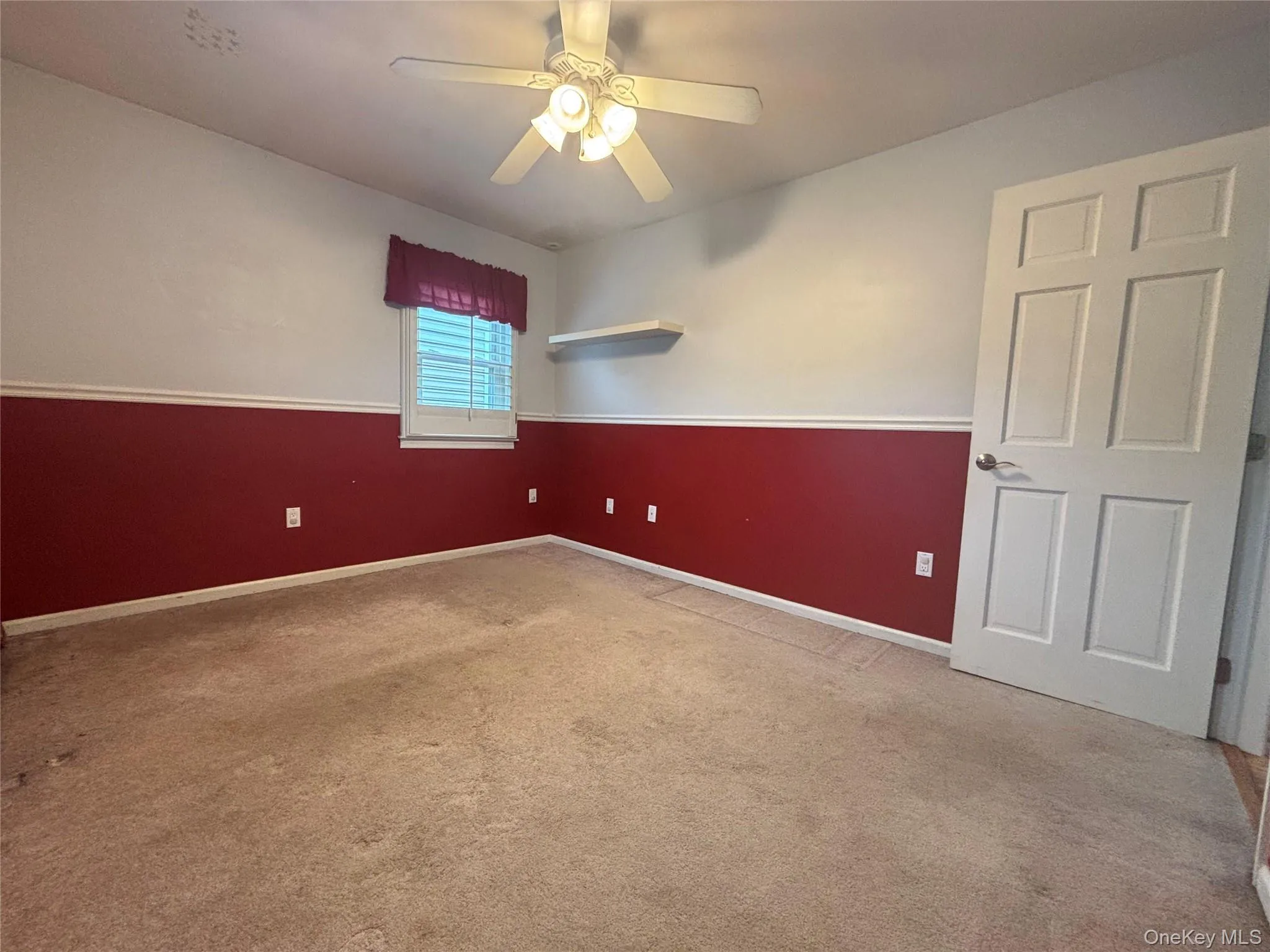Carpeted room featuring a ceiling fan with light fixture, a window with horizontal blinds, a white paneled door, and a wall-mounted shelf Carpeted room featuring a ceiling fan with light fixture, a window with horizontal blinds, a white paneled door, and a wall-mounted shelf