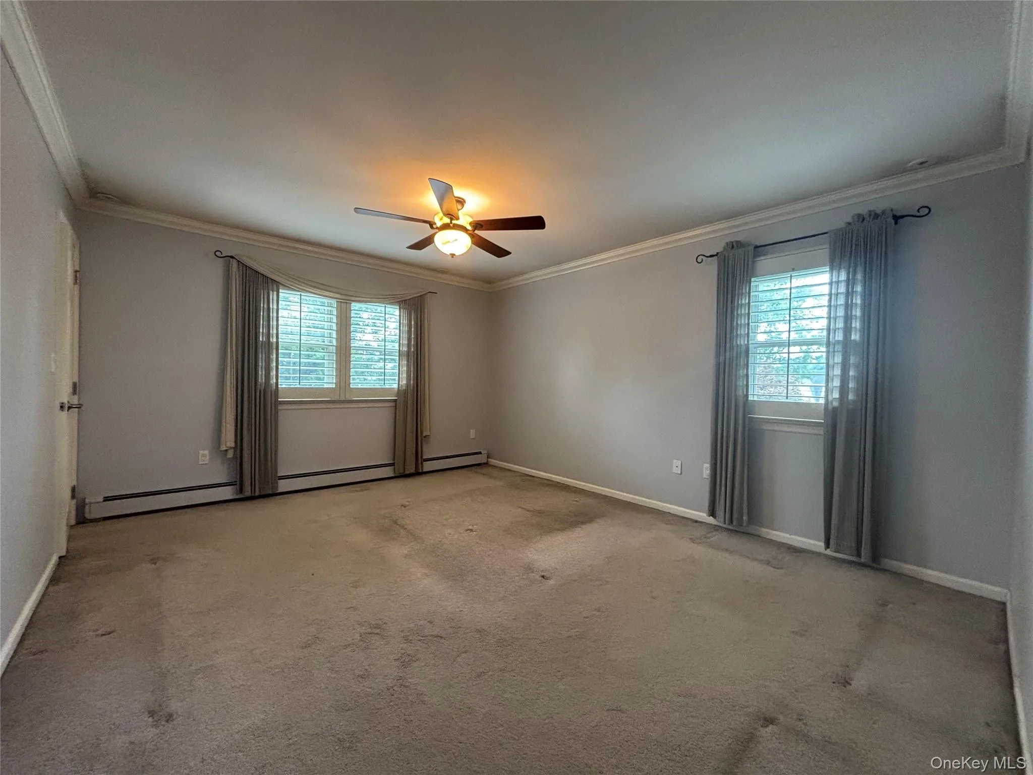 Expansive room featuring two windows with plantation shutters and drapes, crown molding, baseboard trim, and a ceiling fan with integrated lighting Expansive room featuring two windows with plantation shutters and drapes, crown molding, baseboard trim, and a ceiling fan with integrated lighting