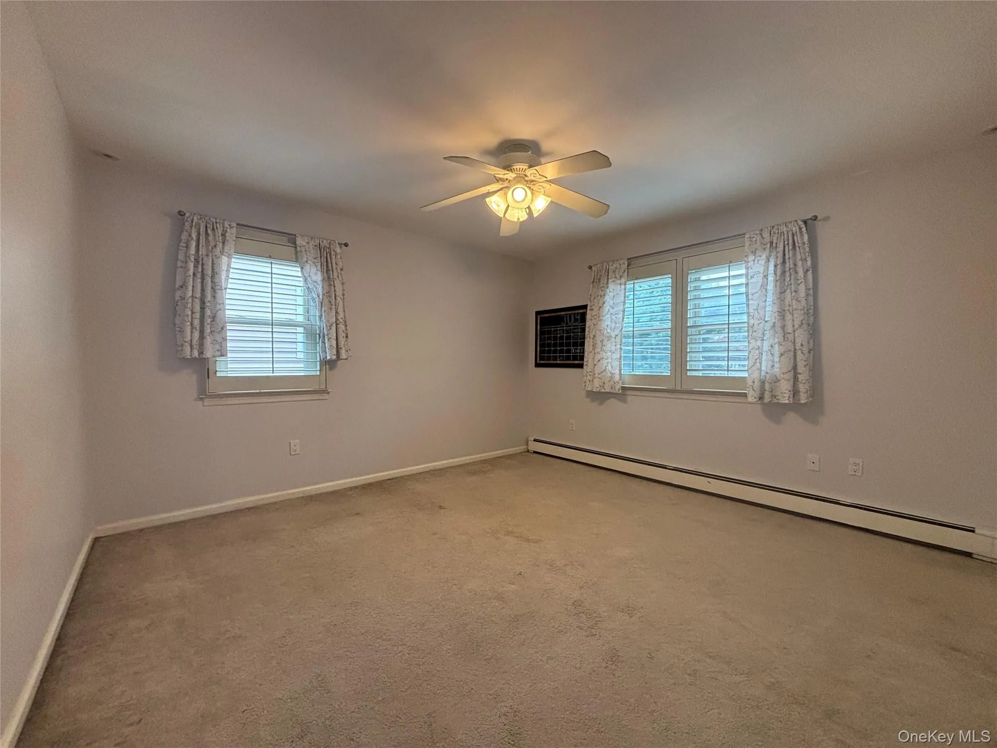 Carpeted room with two windows featuring blinds and drapes, illuminated by a ceiling fan with integrated lighting Carpeted room with two windows featuring blinds and drapes, illuminated by a ceiling fan with integrated lighting