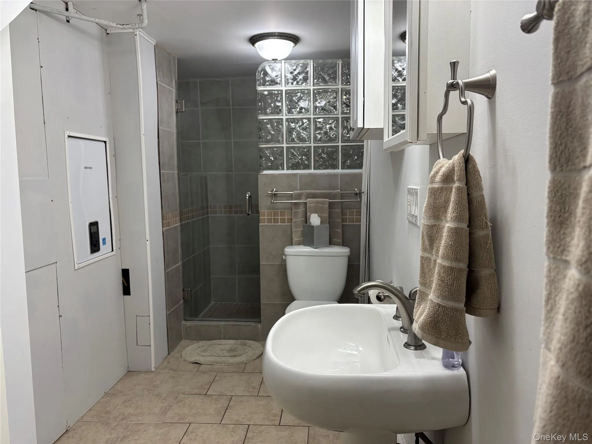 Bathroom featuring a glass-enclosed shower with tile surround, glass block window, pedestal sink with brushed nickel faucet, and tiled flooring Bathroom featuring a glass-enclosed shower with tile surround, glass block window, pedestal sink with brushed nickel faucet, and tiled flooring