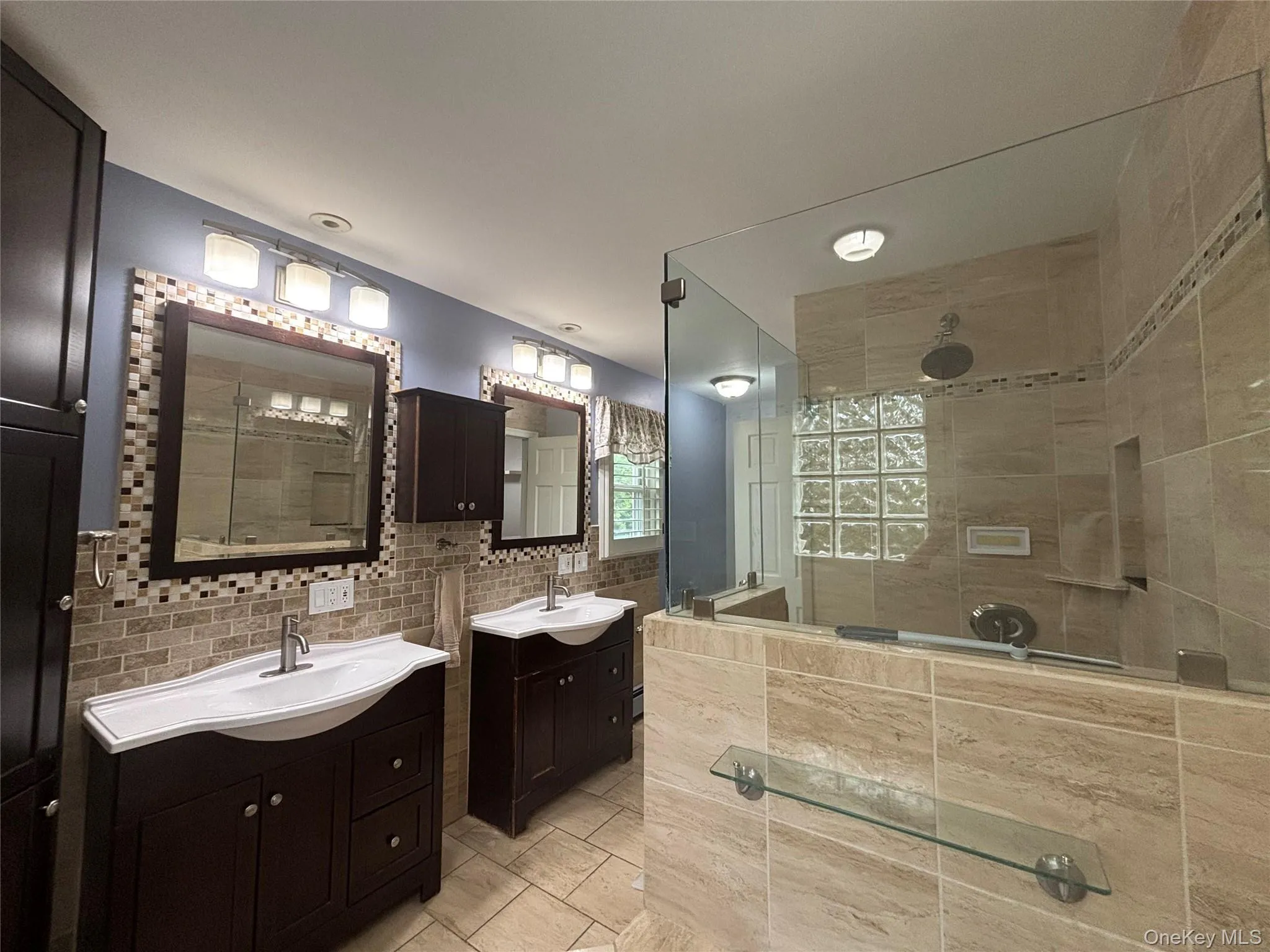 Spacious bathroom featuring a frameless glass shower enclosure, dual vanity sinks with dark wood cabinetry, a tiled shower surround with a built-in niche, and glass block window accents Spacious bathroom featuring a frameless glass shower enclosure, dual vanity sinks with dark wood cabinetry, a tiled shower surround with a built-in niche, and glass block window accents