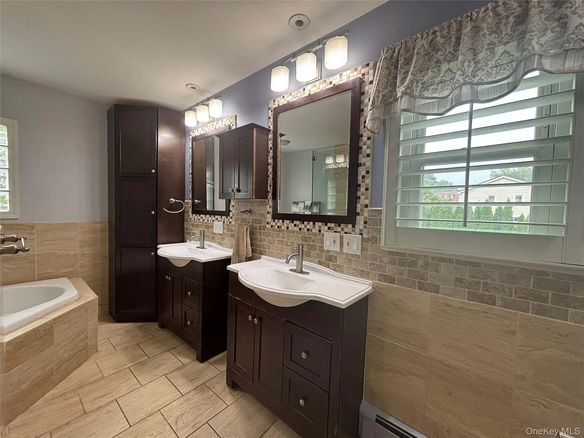 Bathroom featuring dual dark wood-finish vanities with integrated sinks, a built-in soaking tub with tiled surround, full-height storage cabinetry, and tiled flooring extending up the walls Bathroom featuring dual dark wood-finish vanities with integrated sinks, a built-in soaking tub with tiled surround, full-height storage cabinetry, and tiled flooring extending up the walls