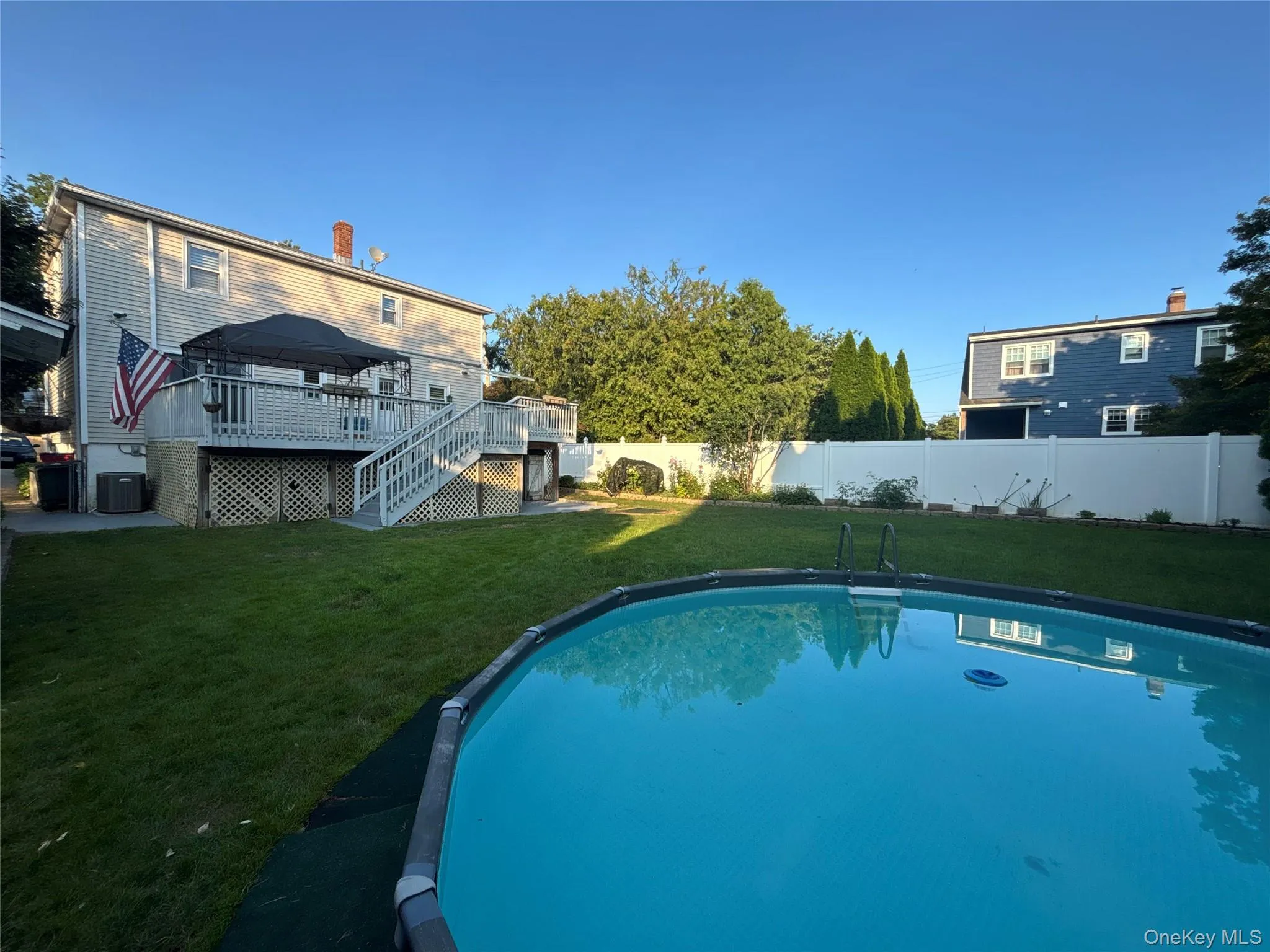 Expansive backyard featuring a sparkling above-ground pool, a spacious multi-level deck, a private white vinyl fence, and mature perimeter landscaping Expansive backyard featuring a sparkling above-ground pool, a spacious multi-level deck, a private white vinyl fence, and mature perimeter landscaping
