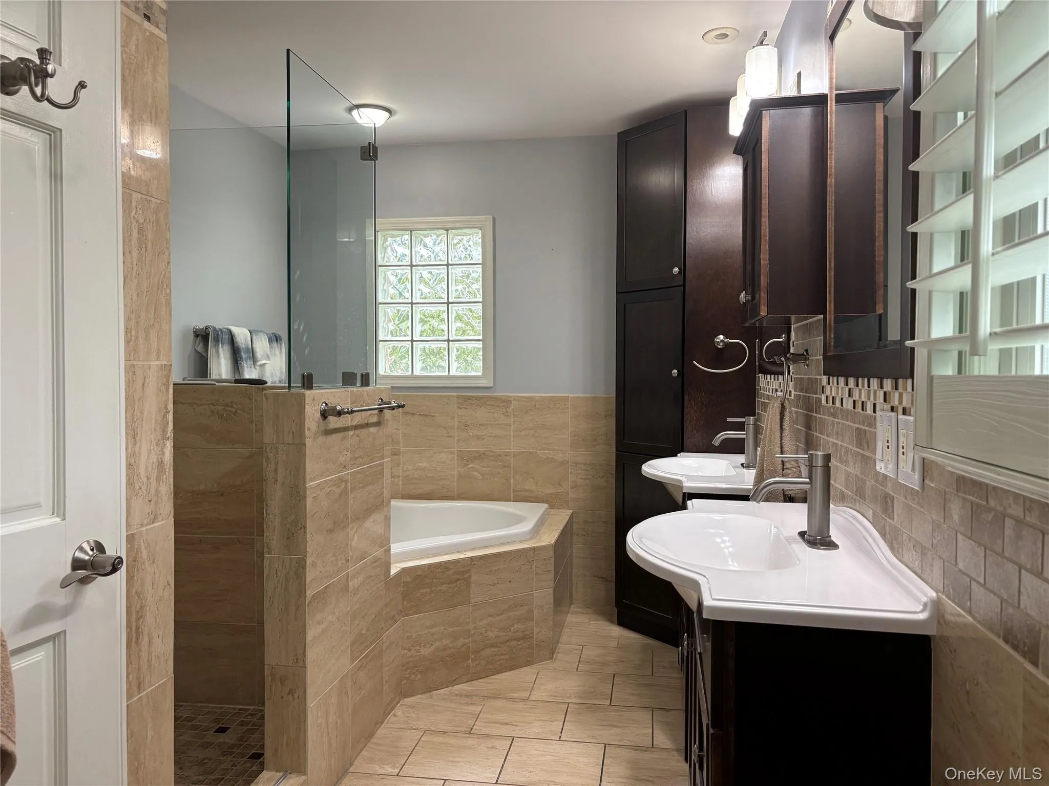 Bathroom featuring dual dark wood vanities with integrated sinks, a tiled backsplash, and a corner soaking tub Bathroom featuring dual dark wood vanities with integrated sinks, a tiled backsplash, and a corner soaking tub