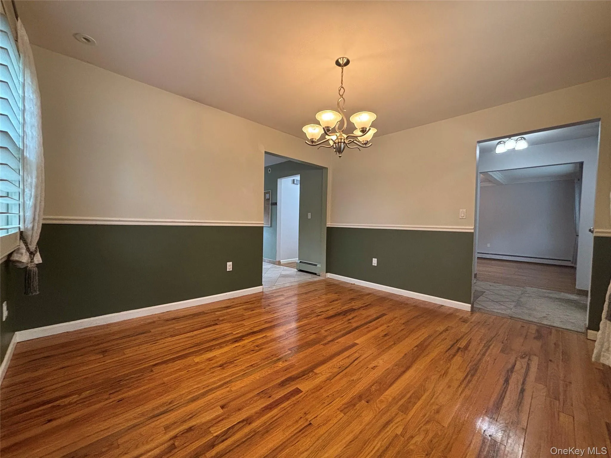 Hardwood flooring throughout, featuring a chandelier with frosted glass shades, a window with plantation shutters, and a two-tone paint scheme with chair rail molding Hardwood flooring throughout, featuring a chandelier with frosted glass shades, a window with plantation shutters, and a two-tone paint scheme with chair rail molding