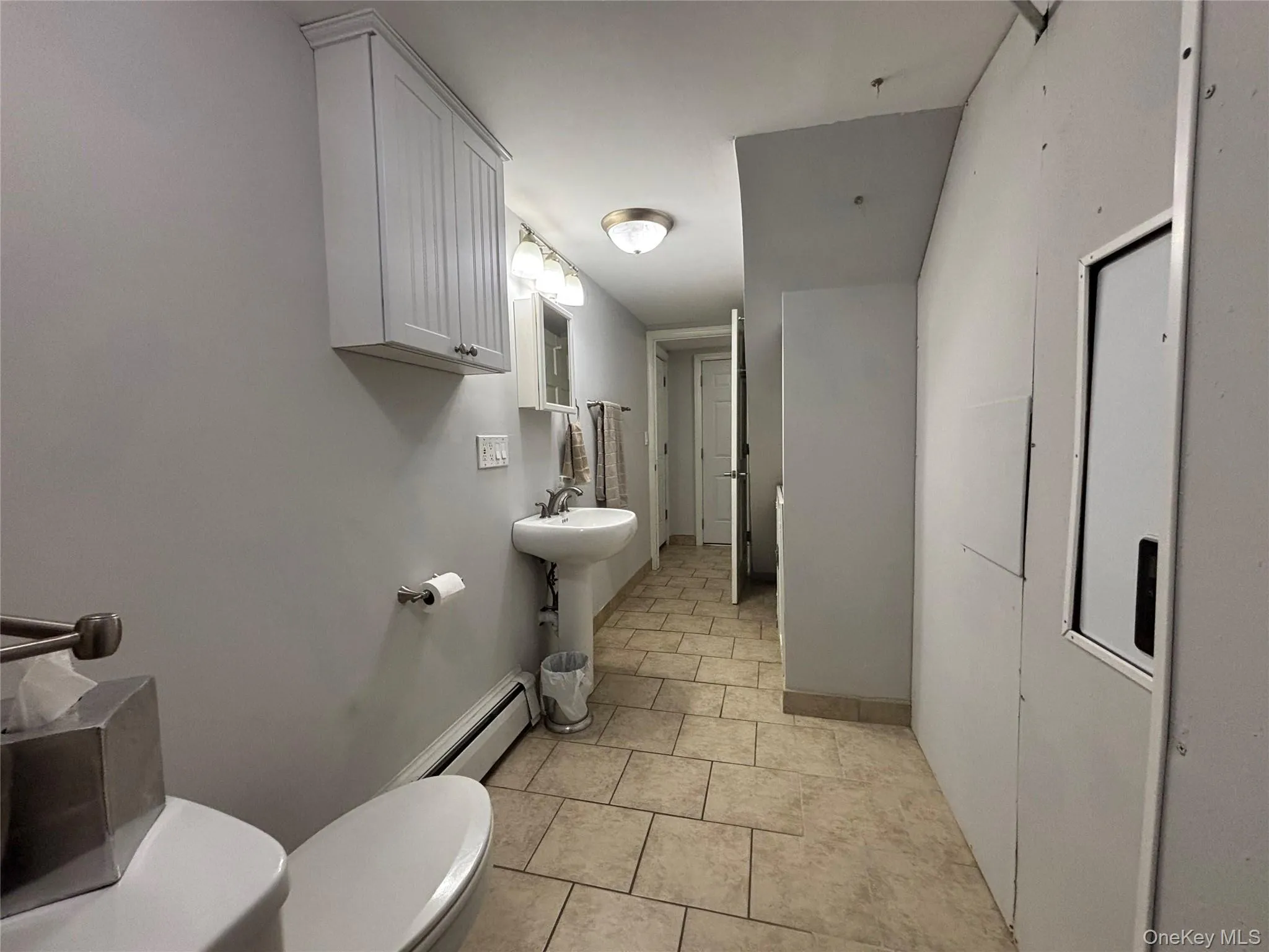 Light-toned tile flooring extends through this bathroom space, featuring a pedestal sink with a wall-mounted light fixture, and a white beadboard-front wall cabinet Light-toned tile flooring extends through this bathroom space, featuring a pedestal sink with a wall-mounted light fixture, and a white beadboard-front wall cabinet