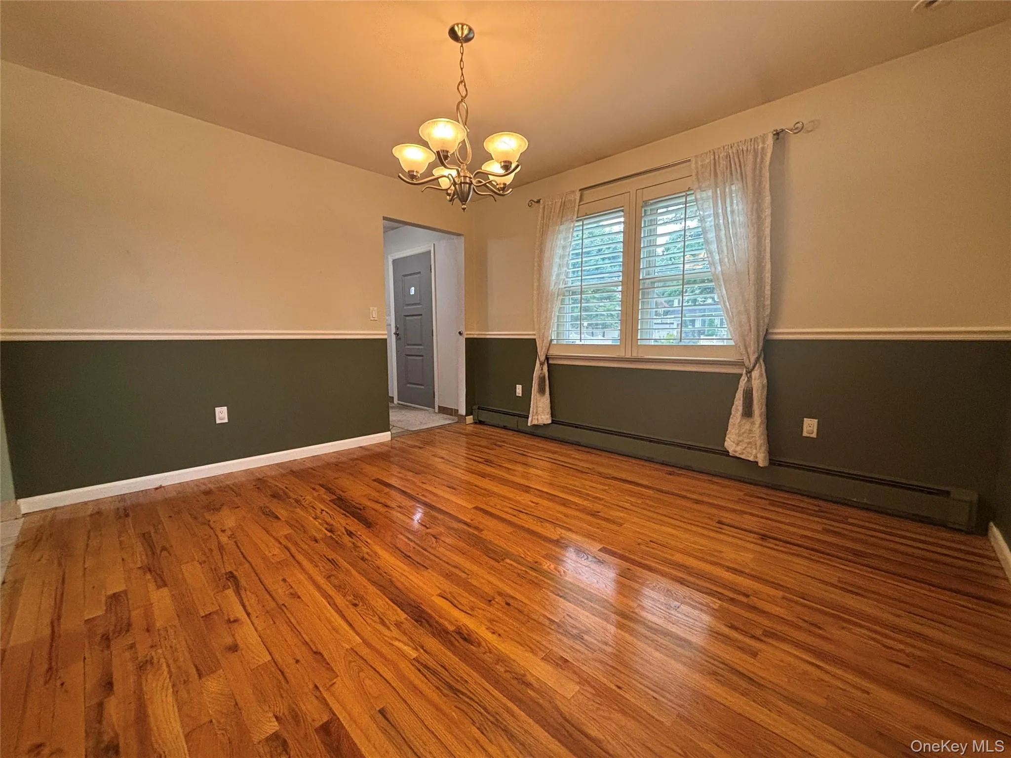 Spacious room featuring rich wood-finish flooring, a contemporary ceiling fixture, a large window with white louvered blinds, and a two-tone wall treatment with chair rail detailing Spacious room featuring rich wood-finish flooring, a contemporary ceiling fixture, a large window with white louvered blinds, and a two-tone wall treatment with chair rail detailing