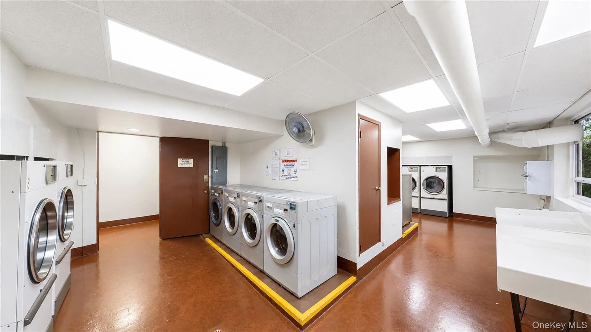 Laundry room featuring multiple front-loading washing machines and dryers, a utility sink, light brown flooring, and bright overhead lighting Laundry room featuring multiple front-loading washing machines and dryers, a utility sink, light brown flooring, and bright overhead lighting