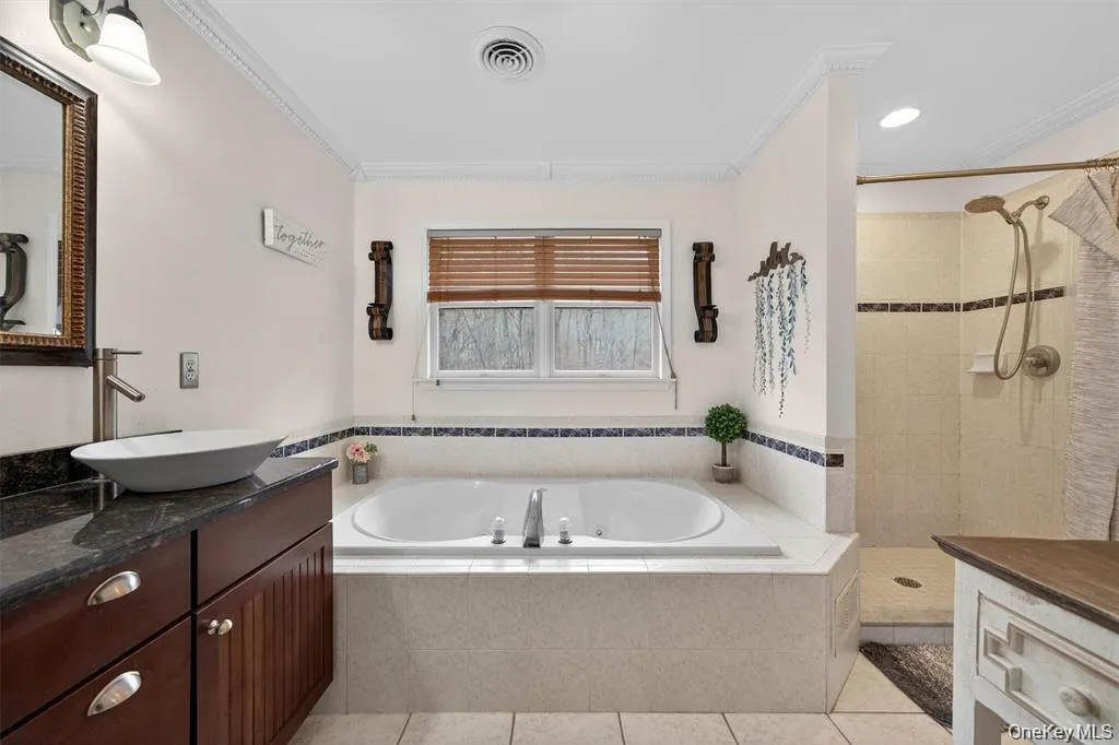 Soaking tub with tiled surround, separate walk-in shower with tiled walls, wood-finish vanity with vessel sink and dark countertop, and decorative crown molding Soaking tub with tiled surround, separate walk-in shower with tiled walls, wood-finish vanity with vessel sink and dark countertop, and decorative crown molding
