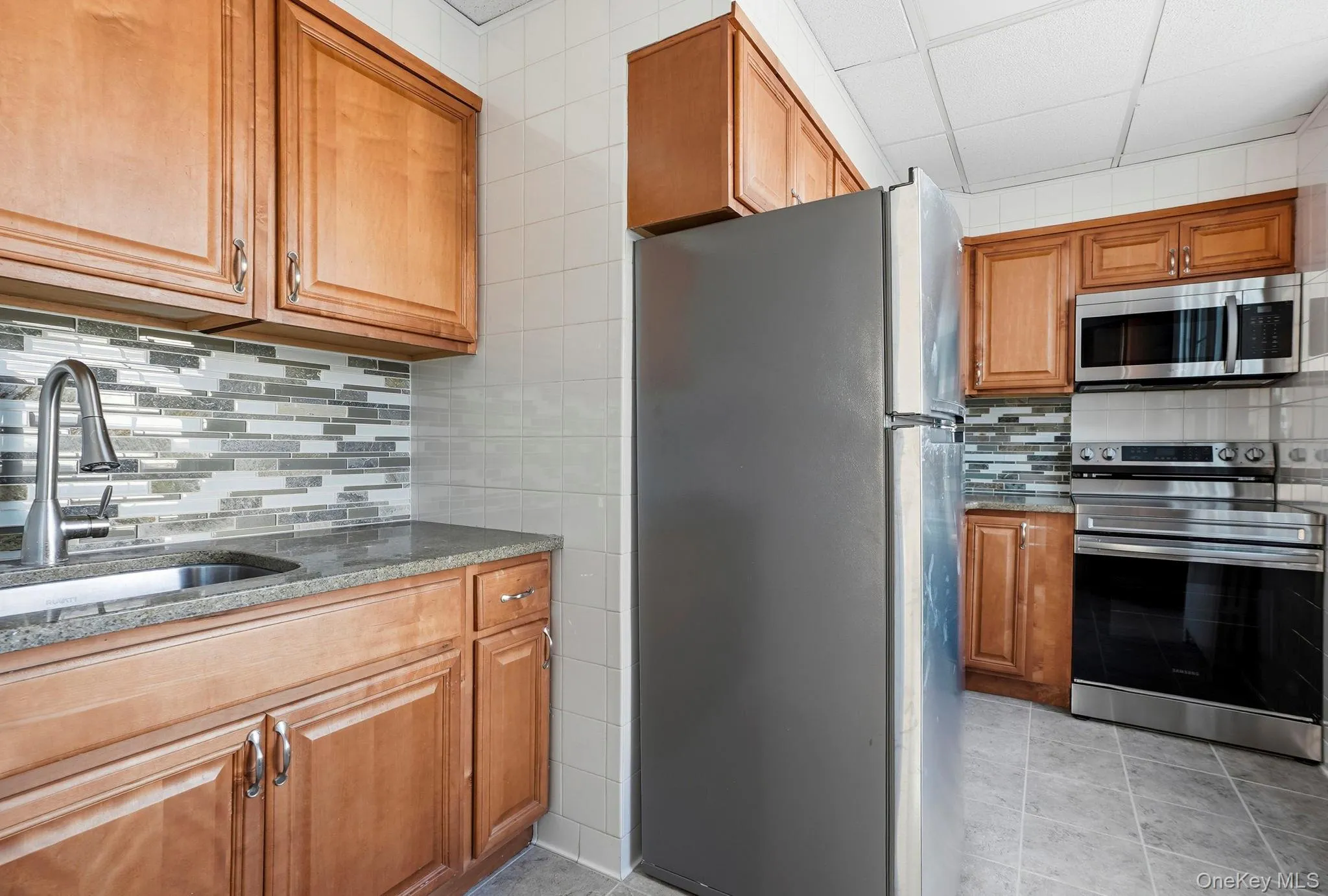 Kitchen featuring wood cabinetry, stainless steel appliances, a multi-tone linear tile backsplash, and stone countertops Kitchen featuring wood cabinetry, stainless steel appliances, a multi-tone linear tile backsplash, and stone countertops