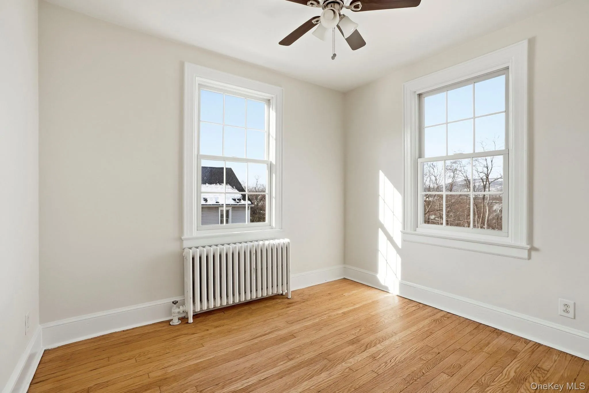 Room featuring wood-finish flooring, two large windows with white trim, a ceiling fan, and a built-in radiator Room featuring wood-finish flooring, two large windows with white trim, a ceiling fan, and a built-in radiator