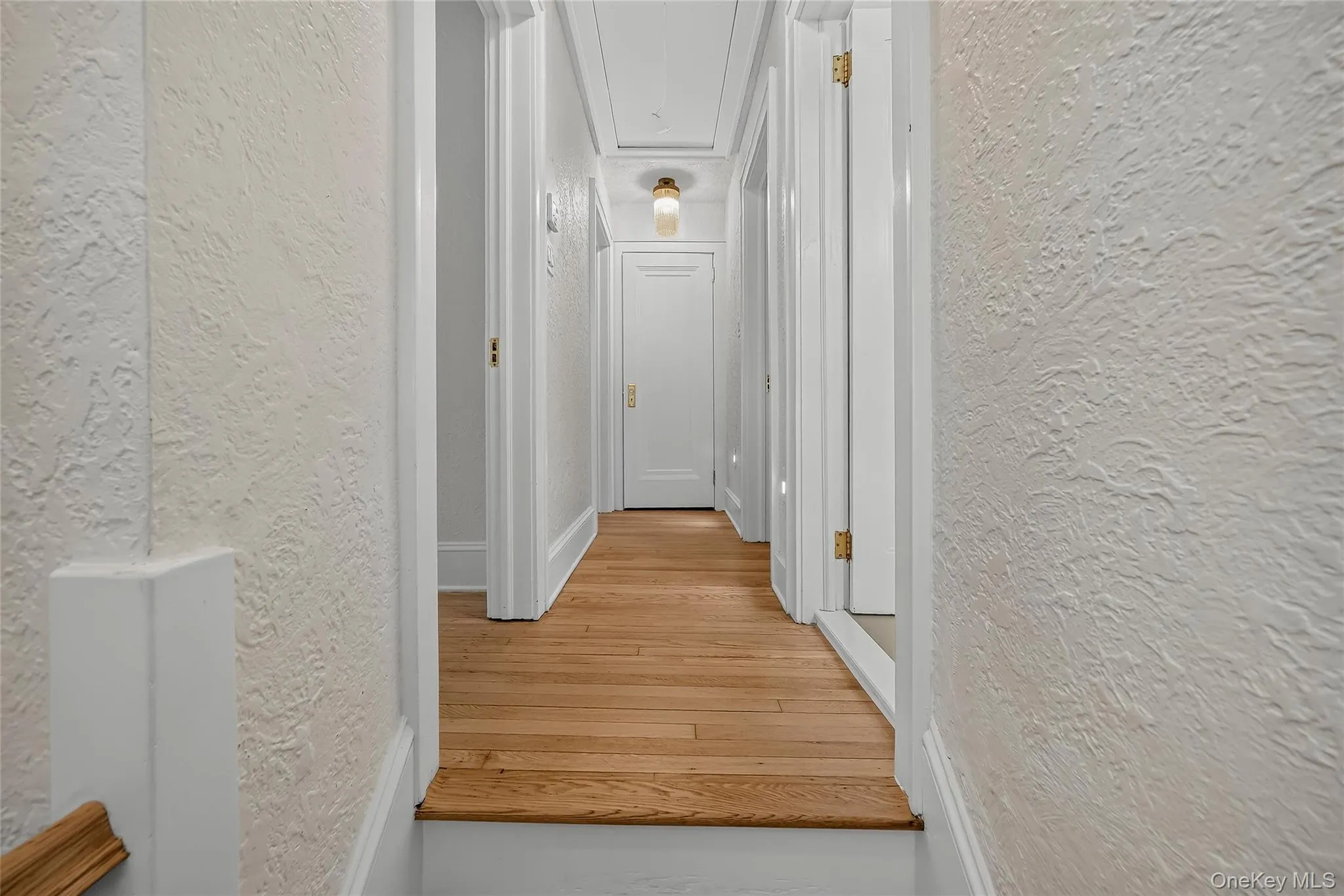 Hallway featuring wood-finish flooring, white doors with brass hardware, textured wall finishes, white trim, and a ceiling-mounted light fixture Hallway featuring wood-finish flooring, white doors with brass hardware, textured wall finishes, white trim, and a ceiling-mounted light fixture