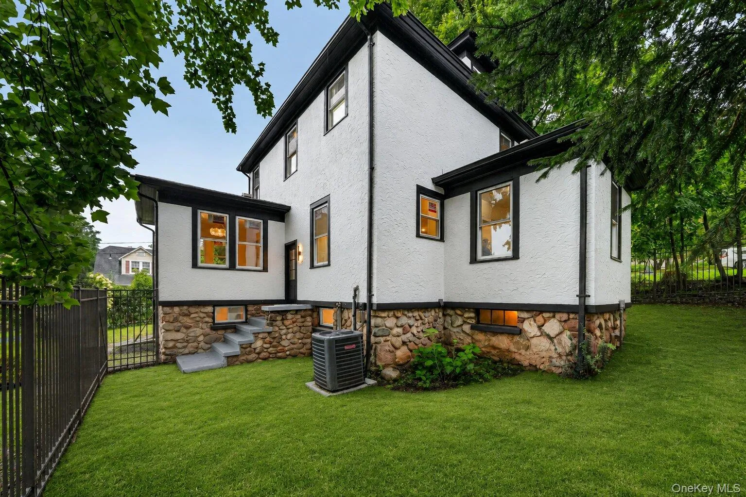 Rear exterior showcasing white stucco siding with black trim, a stone foundation, a manicured lawn, and a black wrought iron fence Rear exterior showcasing white stucco siding with black trim, a stone foundation, a manicured lawn, and a black wrought iron fence
