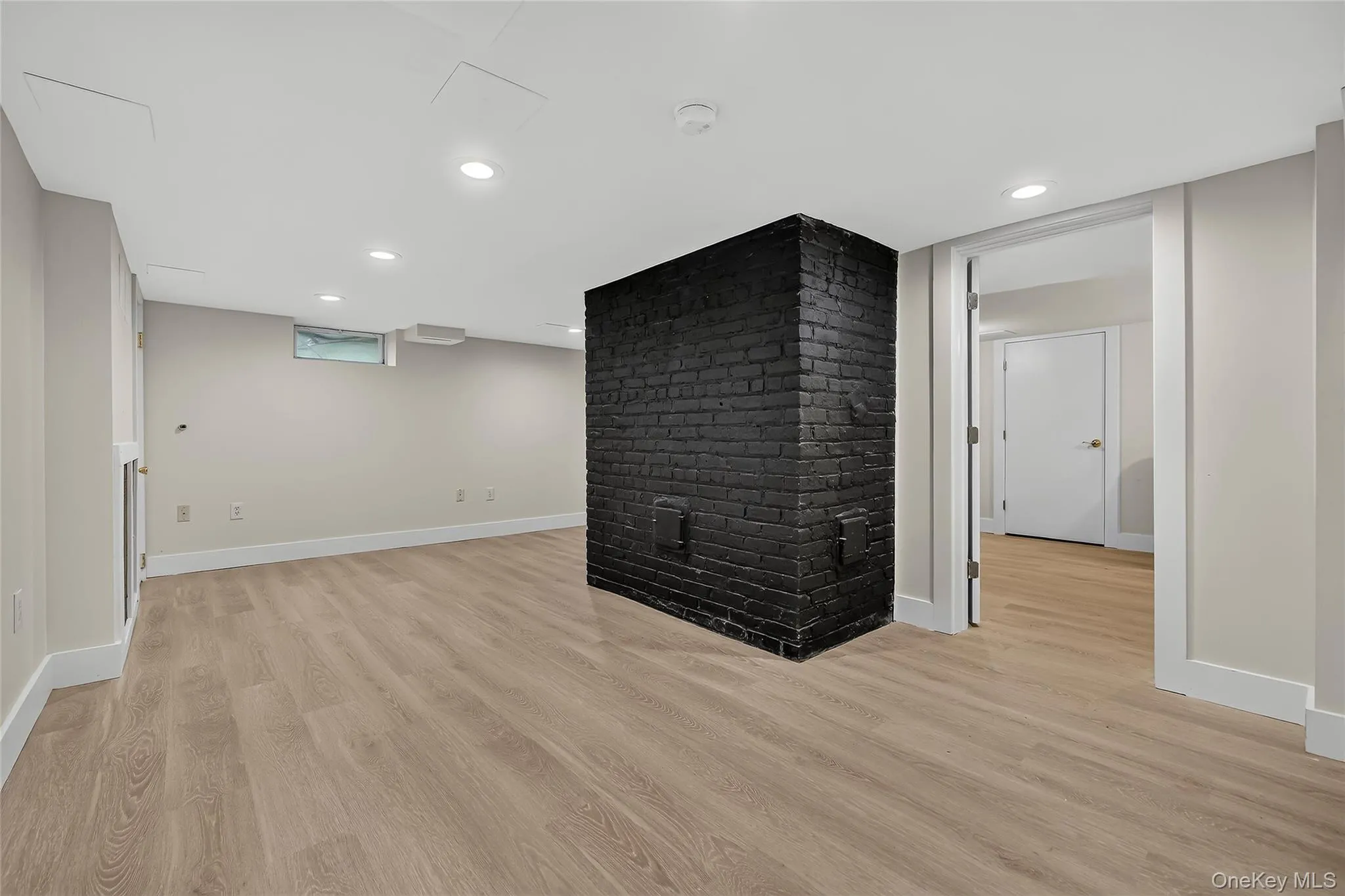 Spacious room with light wood-finish flooring, a painted brick feature, recessed lighting, and a doorway to an additional room with wood-finish flooring Spacious room with light wood-finish flooring, a painted brick feature, recessed lighting, and a doorway to an additional room with wood-finish flooring