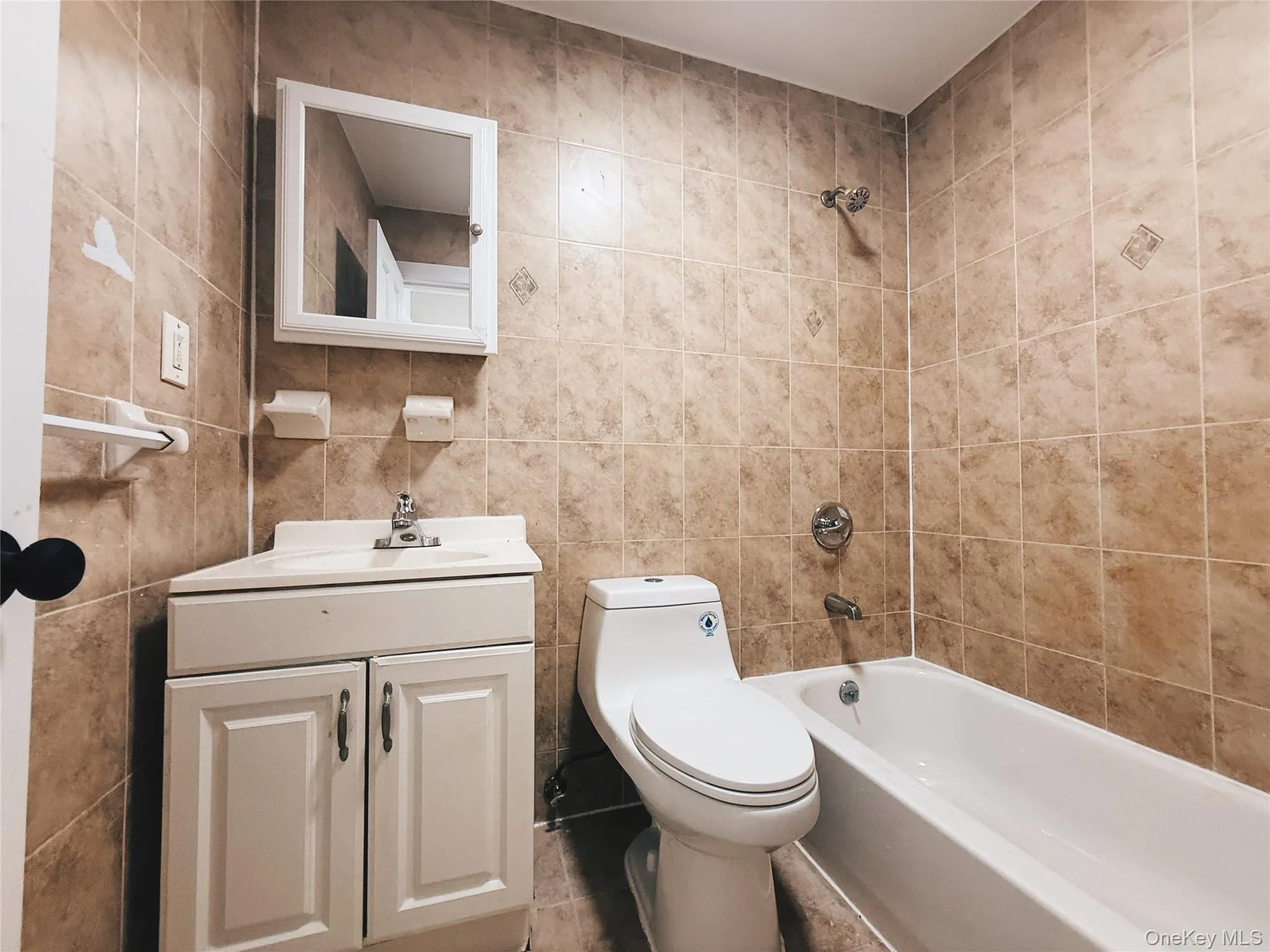 Bathroom featuring full-height tiled walls, white vanity with integrated sink, white medicine cabinet, and a shower-over-tub combination with chrome fixtures. Bathroom featuring full-height tiled walls, white vanity with integrated sink, white medicine cabinet, and a shower-over-tub combination with chrome fixtures.