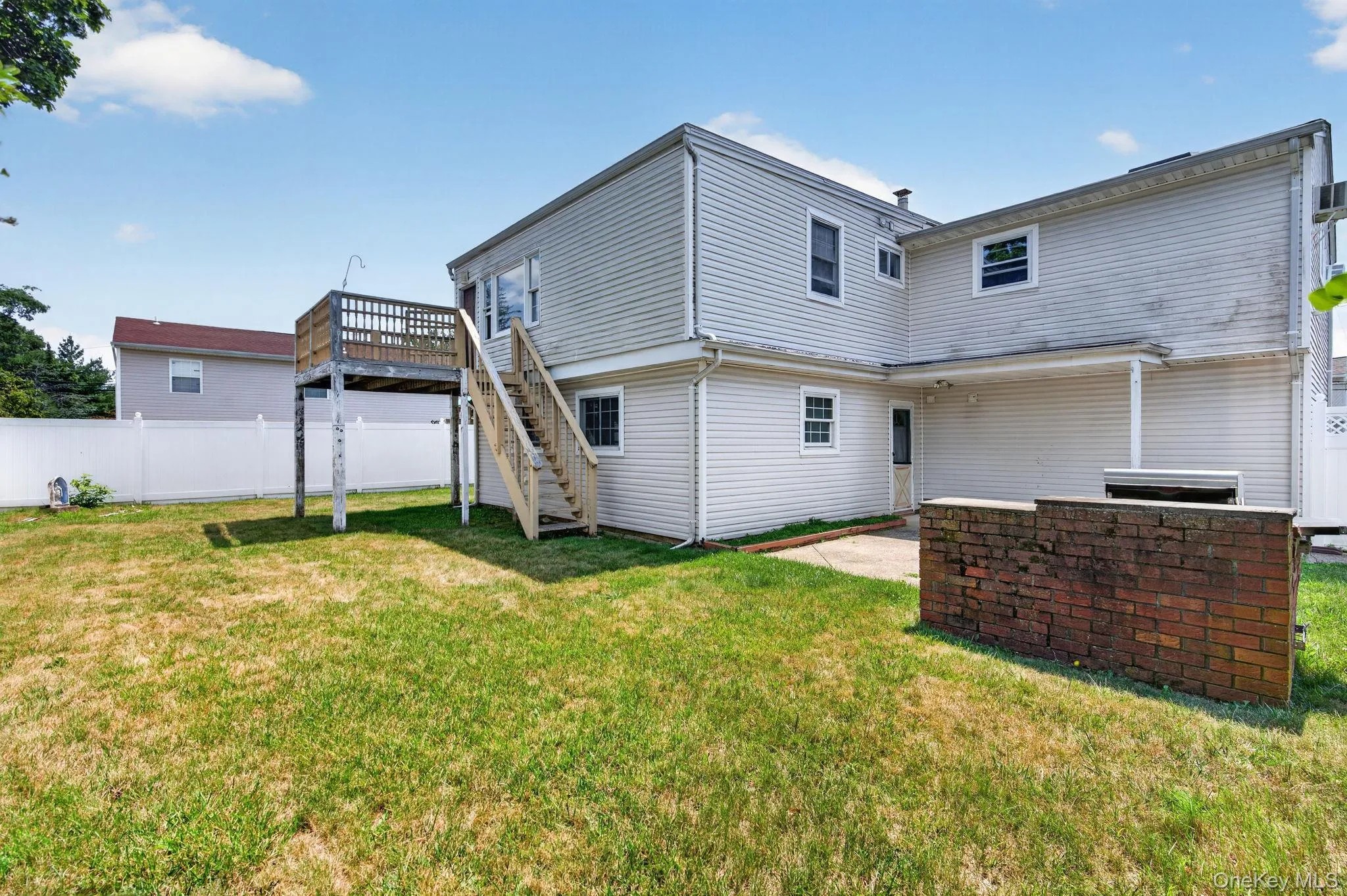 Expansive backyard featuring a concrete patio, a brick-faced outdoor structure, and a raised wooden deck with staircase access Expansive backyard featuring a concrete patio, a brick-faced outdoor structure, and a raised wooden deck with staircase access