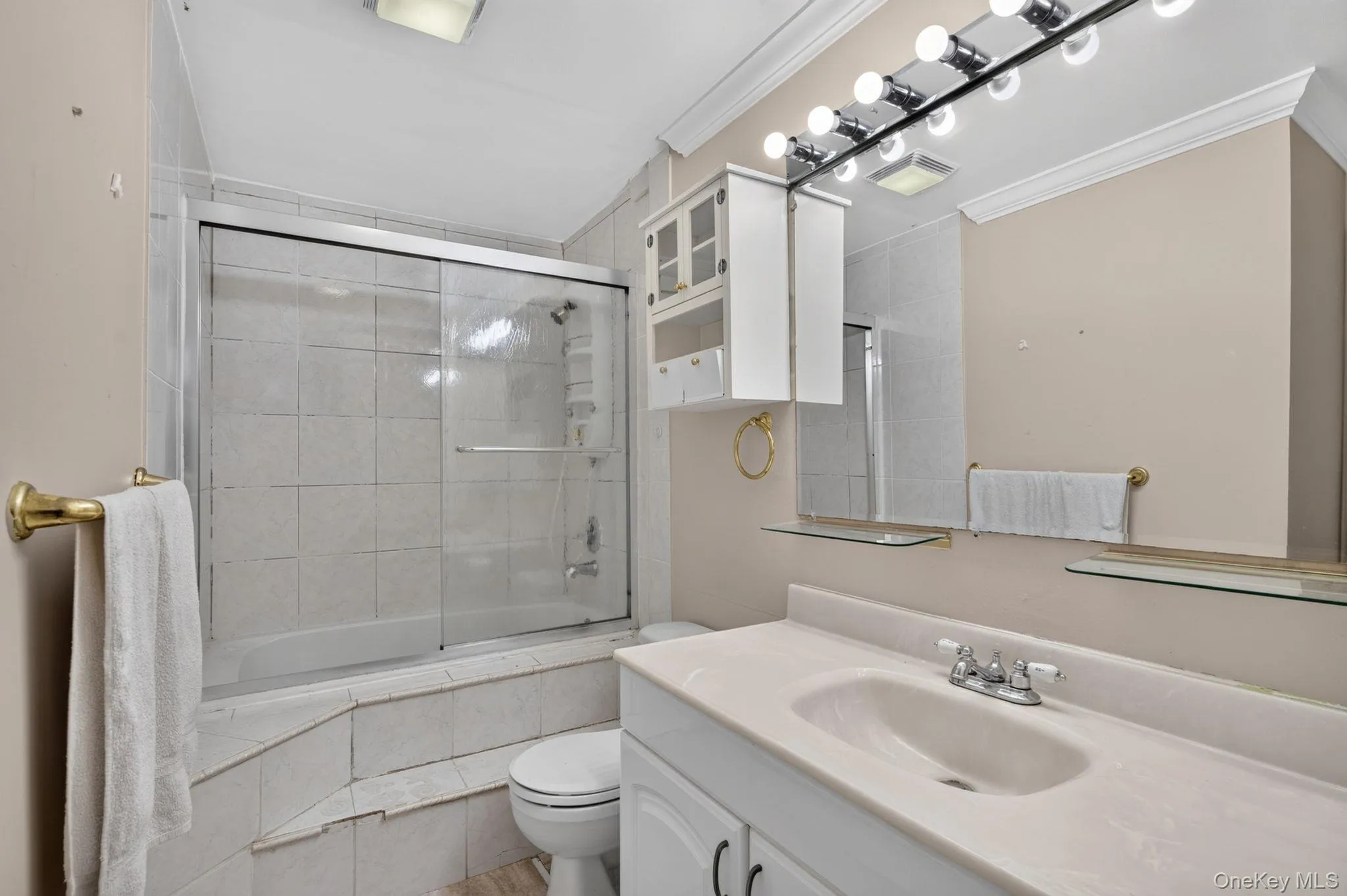 Bathroom featuring a white vanity with integrated sink, a mirrored wall with strip lighting, built-in wall cabinets, and a bathtub with sliding glass doors Bathroom featuring a white vanity with integrated sink, a mirrored wall with strip lighting, built-in wall cabinets, and a bathtub with sliding glass doors