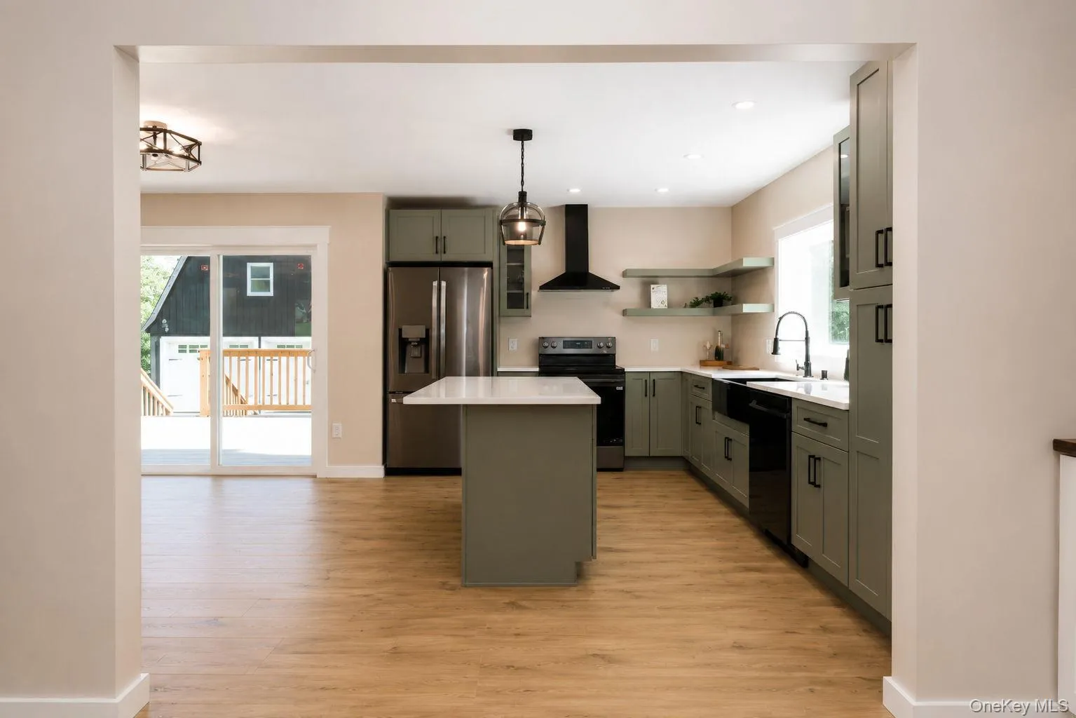 Modern kitchen featuring wood-finish flooring, a central island with a white countertop, sage-toned cabinetry, stainless steel appliances, and a black range hood Modern kitchen featuring wood-finish flooring, a central island with a white countertop, sage-toned cabinetry, stainless steel appliances, and a black range hood