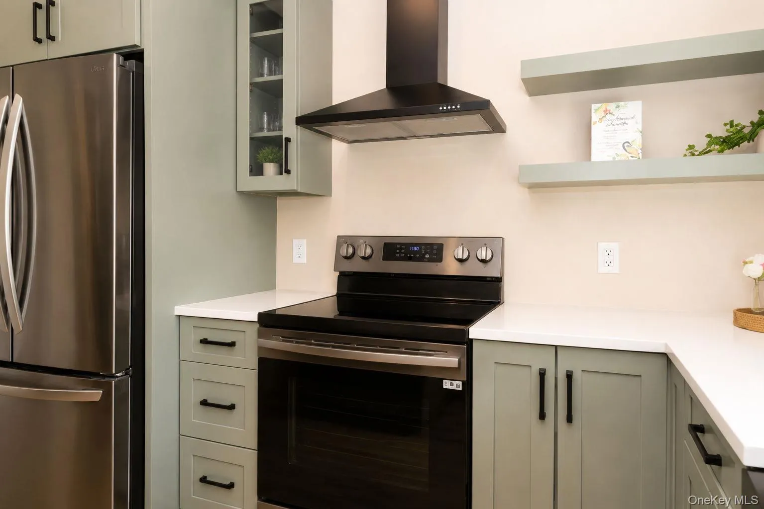 Modern kitchen featuring a stainless steel refrigerator, contemporary range with a black vent hood, light green cabinetry, white countertops, and open shelving Modern kitchen featuring a stainless steel refrigerator, contemporary range with a black vent hood, light green cabinetry, white countertops, and open shelving
