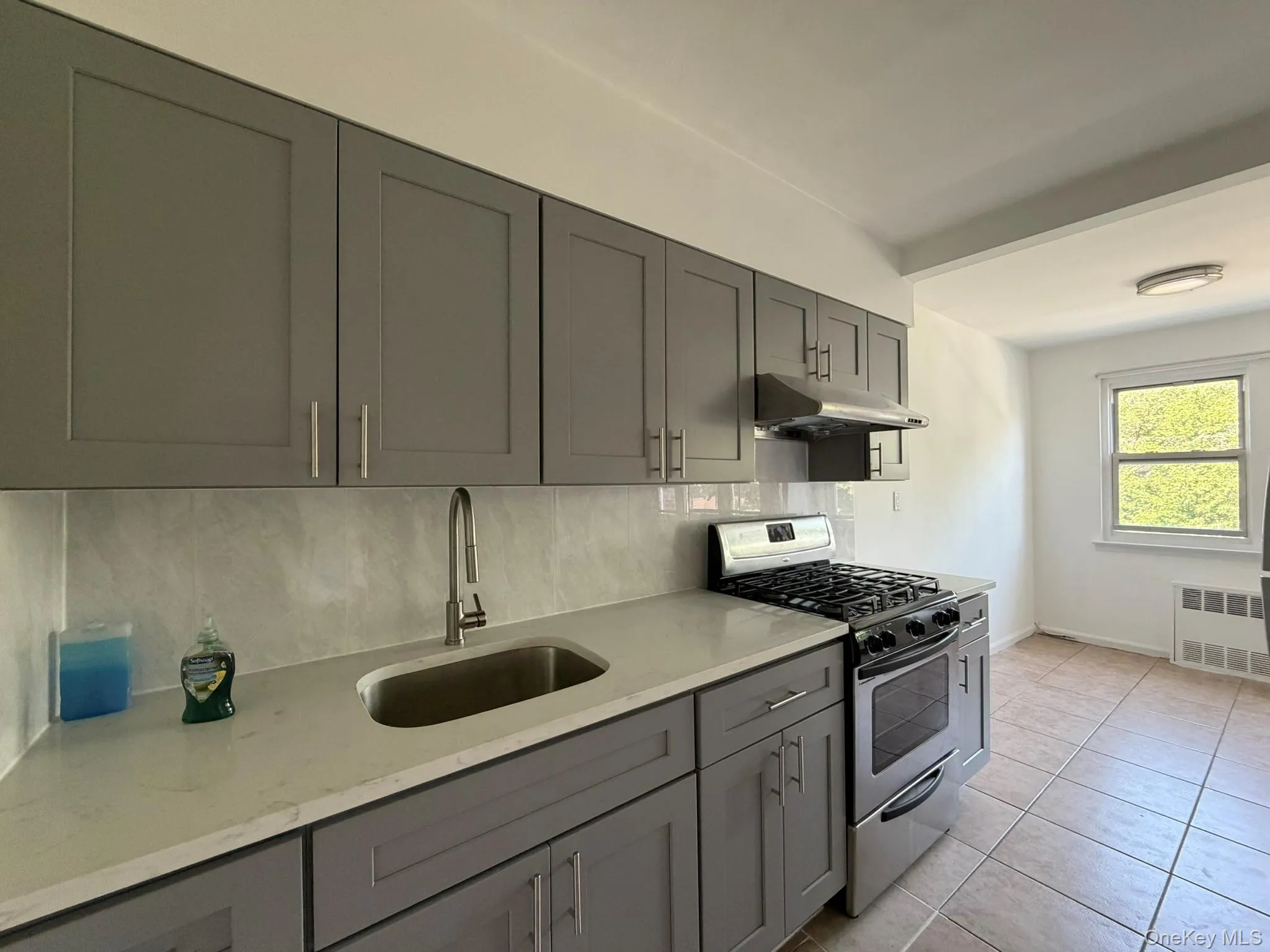 Modern kitchen featuring gray shaker-style cabinetry with brushed nickel hardware, light-toned countertops, and a stainless steel gas range with overhead ventilation Modern kitchen featuring gray shaker-style cabinetry with brushed nickel hardware, light-toned countertops, and a stainless steel gas range with overhead ventilation