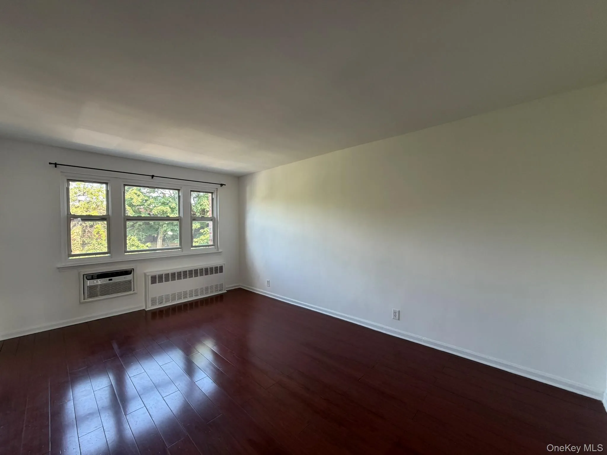 Interior room featuring dark wood-finish flooring, white walls, and a large window with three panes Interior room featuring dark wood-finish flooring, white walls, and a large window with three panes