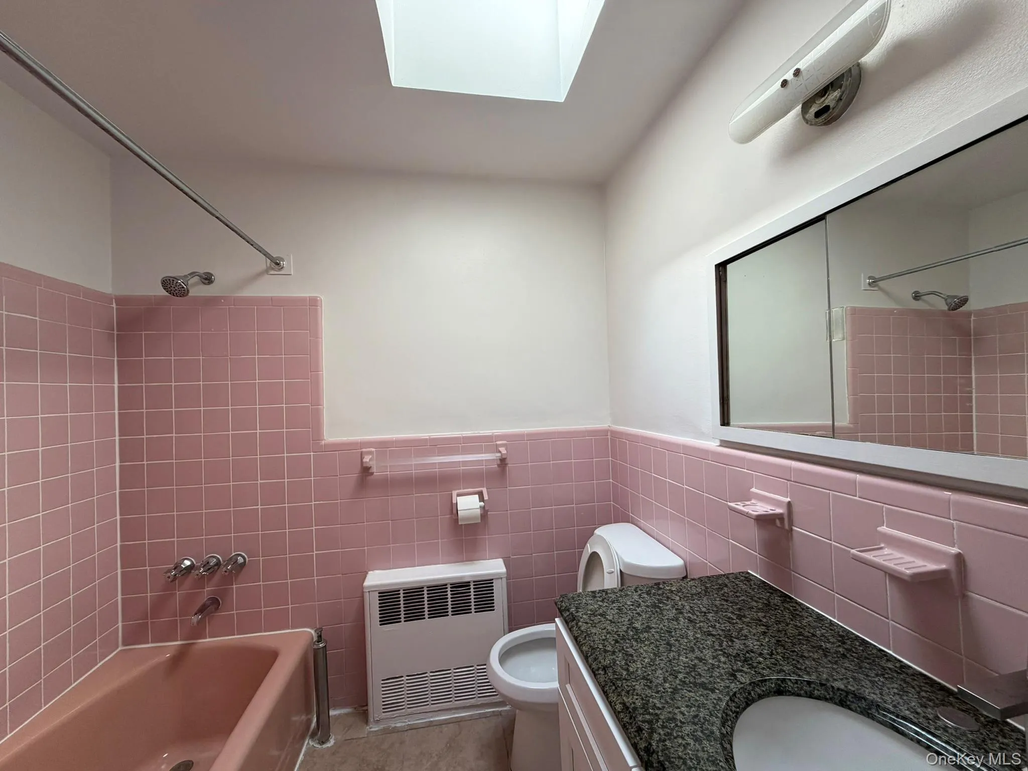 Bathroom featuring pink tile walls, a matching pink bathtub, a skylight, a vanity with a dark speckled countertop and integrated sink, and a mirrored medicine cabinet Bathroom featuring pink tile walls, a matching pink bathtub, a skylight, a vanity with a dark speckled countertop and integrated sink, and a mirrored medicine cabinet