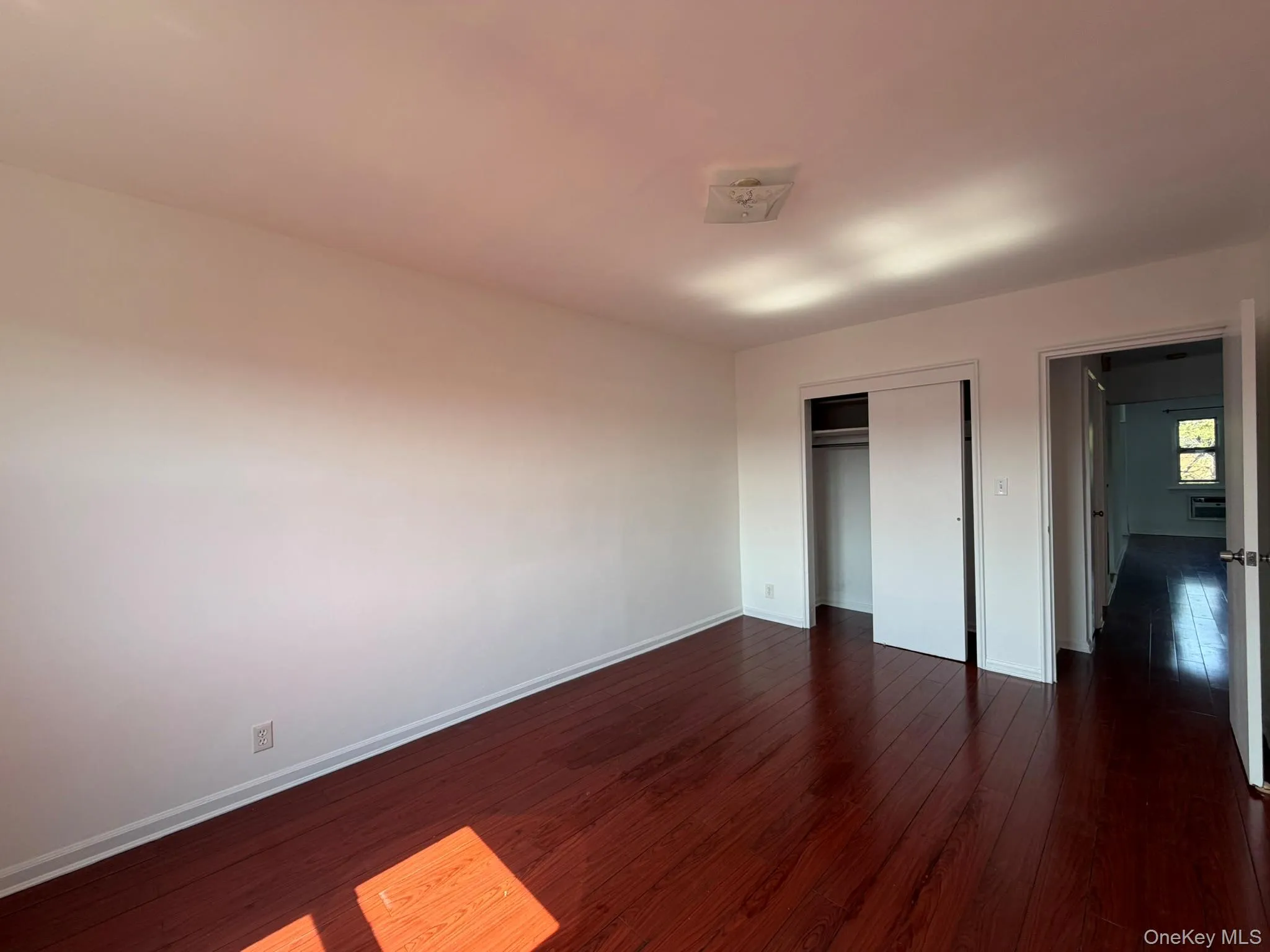 Spacious room featuring rich wood-finish flooring, white walls, and a ceiling-mounted light fixture Spacious room featuring rich wood-finish flooring, white walls, and a ceiling-mounted light fixture