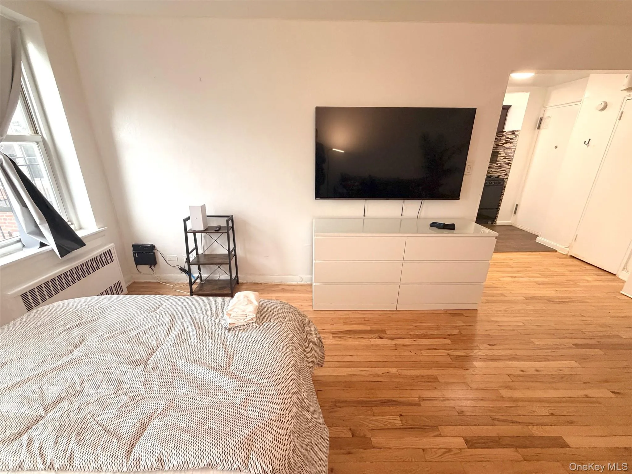 Hardwood flooring paired with white walls, featuring a window with a radiator below, built-in wall-mounted television, and a white dresser Hardwood flooring paired with white walls, featuring a window with a radiator below, built-in wall-mounted television, and a white dresser