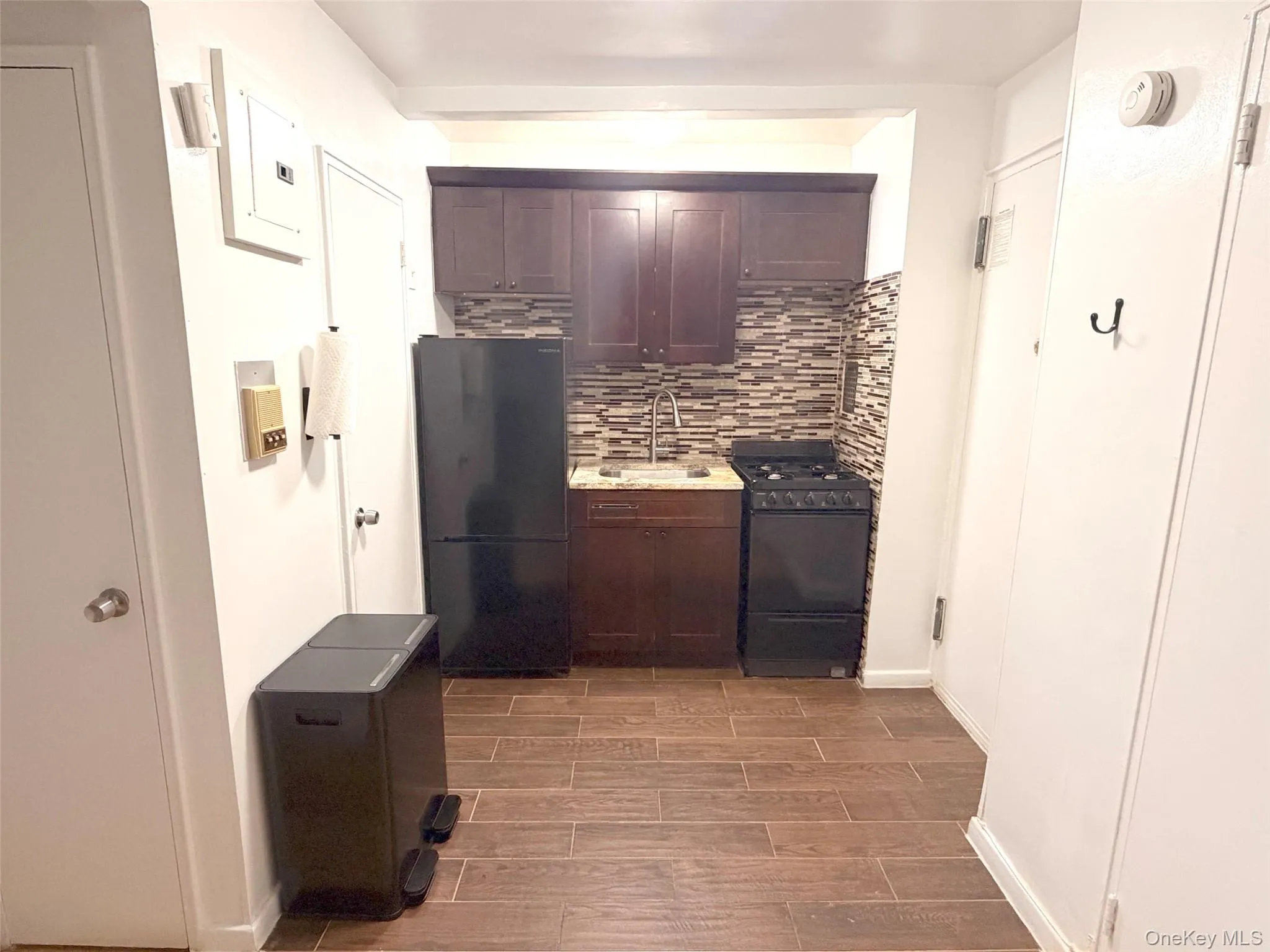Compact kitchen featuring a mosaic tile backsplash, dark wood-finish cabinetry, stainless steel sink with gooseneck faucet, black refrigerator, and black range Compact kitchen featuring a mosaic tile backsplash, dark wood-finish cabinetry, stainless steel sink with gooseneck faucet, black refrigerator, and black range