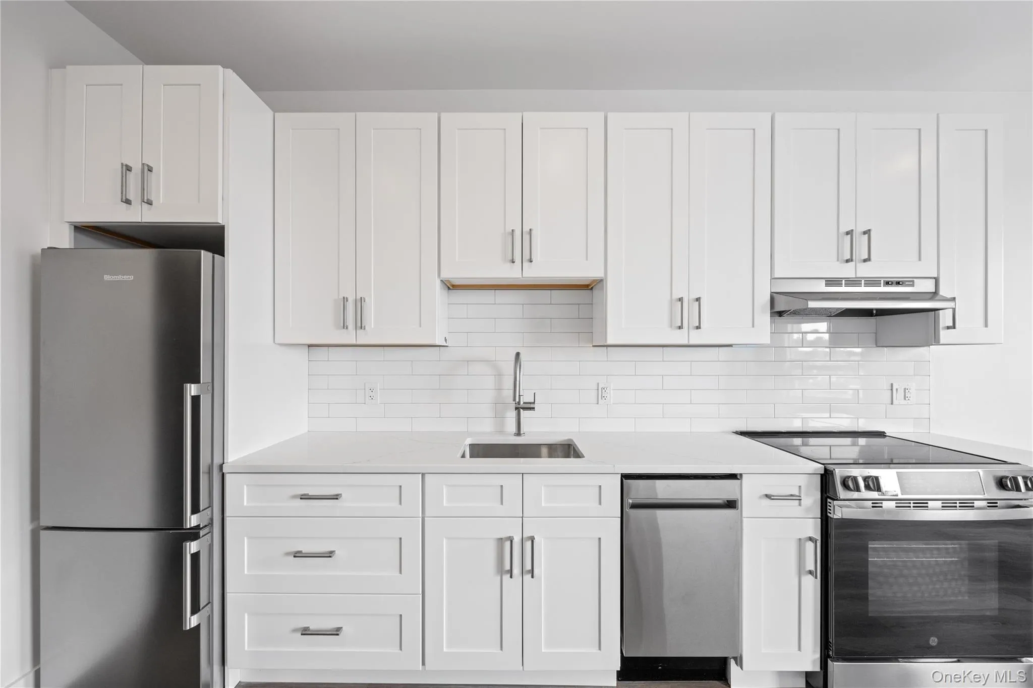 Modern kitchen featuring white shaker cabinetry, stainless steel appliances, a subway tile backsplash, light-toned countertops, and a stainless steel sink with a gooseneck faucet Modern kitchen featuring white shaker cabinetry, stainless steel appliances, a subway tile backsplash, light-toned countertops, and a stainless steel sink with a gooseneck faucet