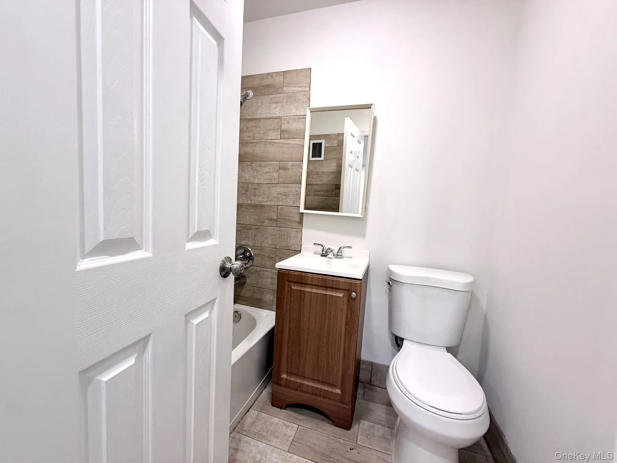 Bathroom featuring a white paneled door, wood-finish tile flooring, a white vanity with wood-finish cabinet, a white toilet, and a tiled shower surround Bathroom featuring a white paneled door, wood-finish tile flooring, a white vanity with wood-finish cabinet, a white toilet, and a tiled shower surround