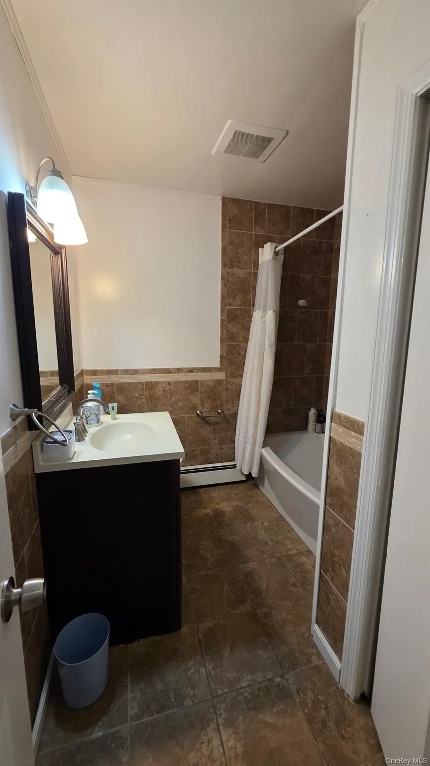 Bathroom featuring a vanity with an integrated sink, a rectangular mirror, a bathtub with a shower, and floor-to-ceiling tile work Bathroom featuring a vanity with an integrated sink, a rectangular mirror, a bathtub with a shower, and floor-to-ceiling tile work