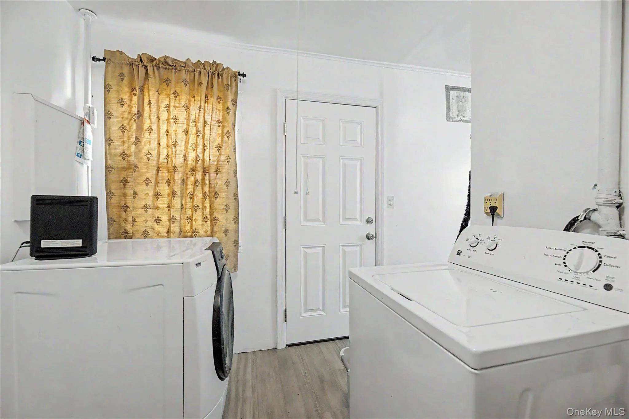 Dedicated laundry room featuring side-by-side front-load washer and dryer with white cabinetry, wood-finish flooring, natural light from a window with yellow curtains, and direct access to the home's interior spaces Dedicated laundry room featuring side-by-side front-load washer and dryer with white cabinetry, wood-finish flooring, natural light from a window with yellow curtains, and direct access to the home's interior spaces