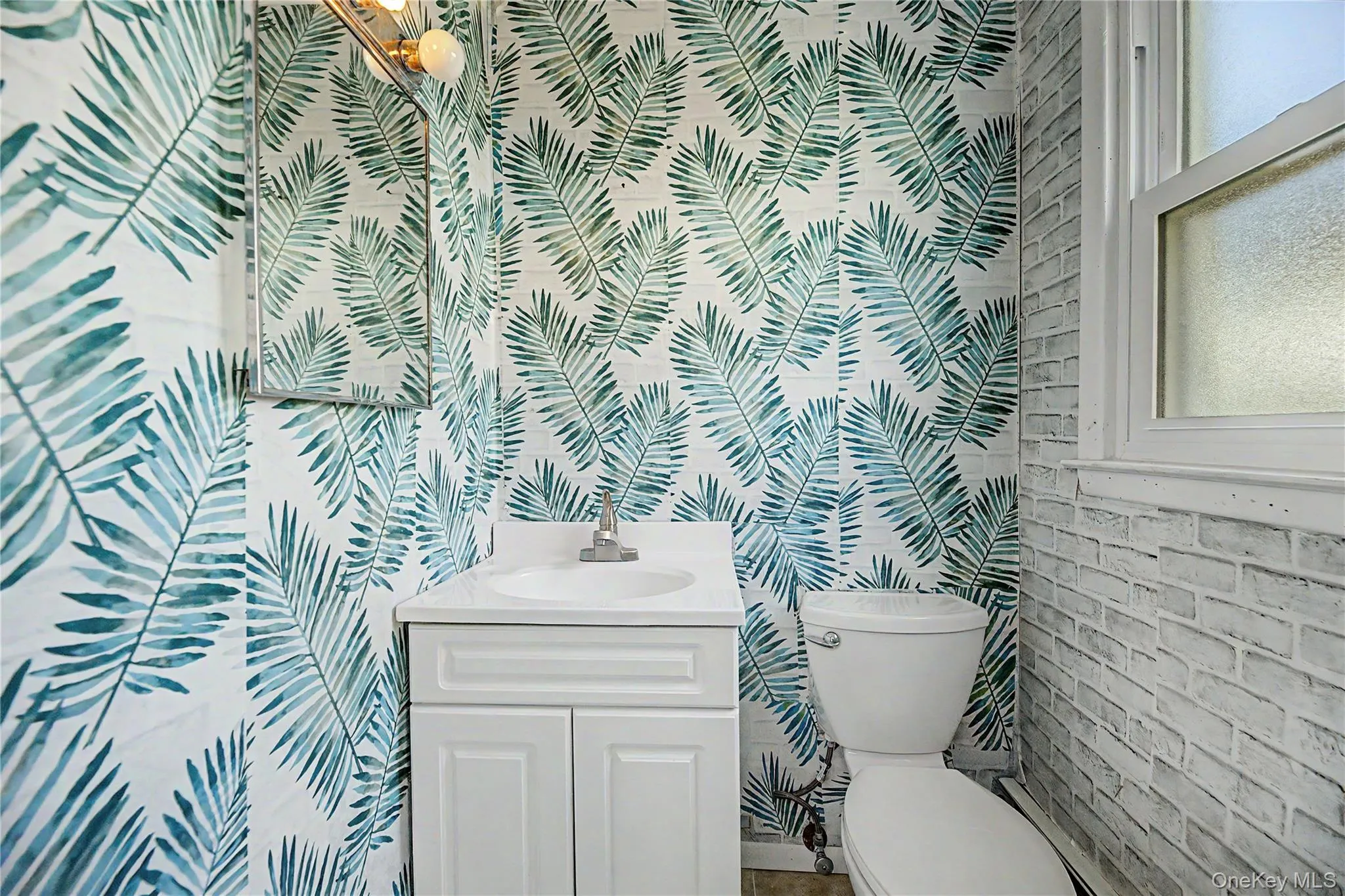 Powder room featuring botanical-patterned wallcovering in soft green and blue tones, white cabinetry with integrated sink vanity, ceramic toilet, and brick-textured accent wall Powder room featuring botanical-patterned wallcovering in soft green and blue tones, white cabinetry with integrated sink vanity, ceramic toilet, and brick-textured accent wall