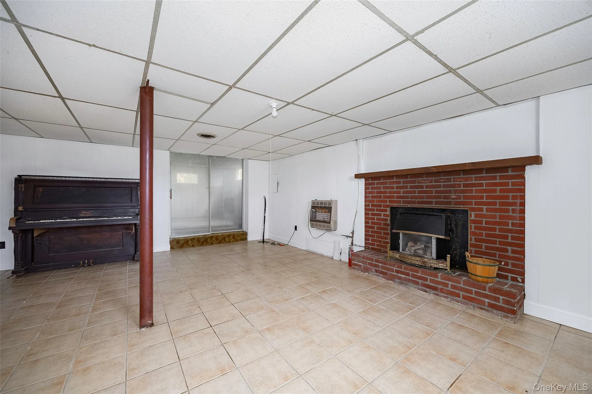 Finished below grade area with a brick fireplace, a drop ceiling, light tile patterned flooring, and heating unit Finished below grade area with a brick fireplace, a drop ceiling, light tile patterned flooring, and heating unit